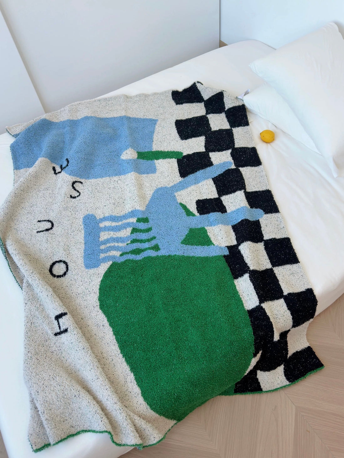 A textured knit throw blanket with a colorful abstract design and a black-and-white checkered border, draped over a white bed. The design features green, light blue, and gray blocks with the word 'HOUSE' printed above.