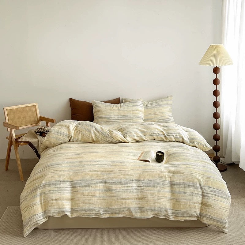 A bed dressed in a cream and beige striped bedding set with matching pillows, styled in a minimalist bedroom with a wooden chair, floor lamp, and light-colored walls.