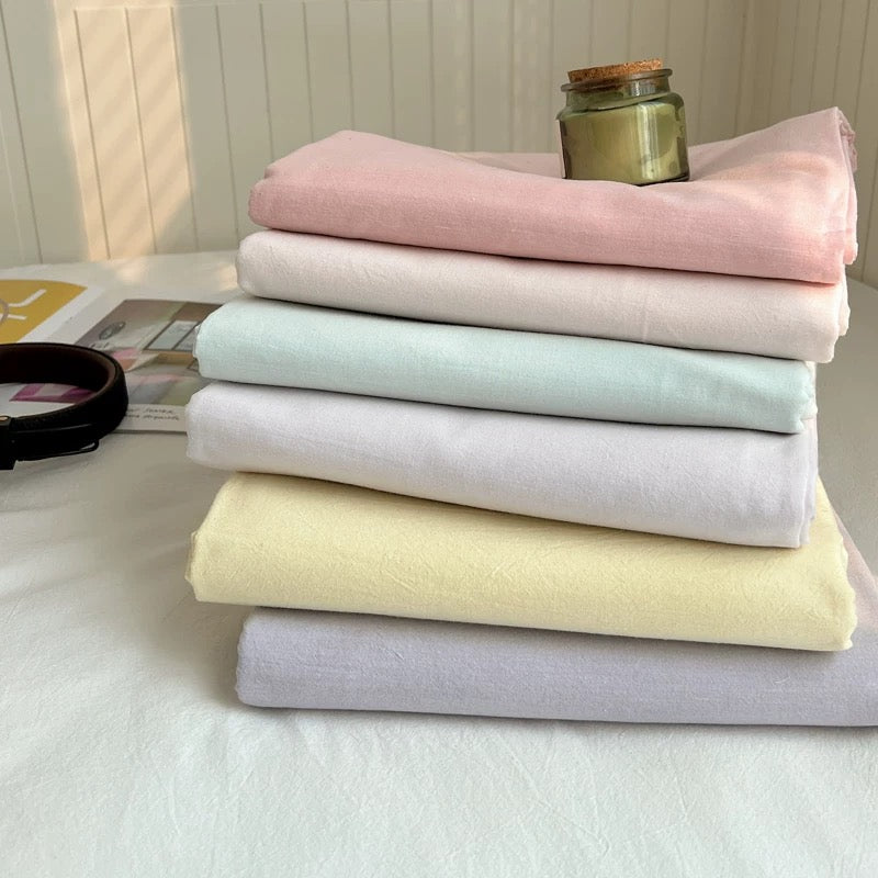 A stack of six folded cotton fabrics in pastel shades of pink, cream, mint, light purple, pale yellow, and lavender, arranged on a white surface with a small glass jar on top.