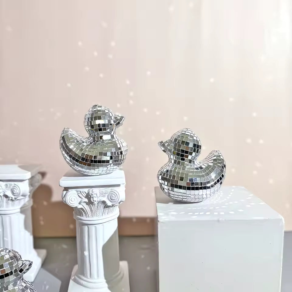 Two silver disco duck decorations on white pedestals with a soft pink background.