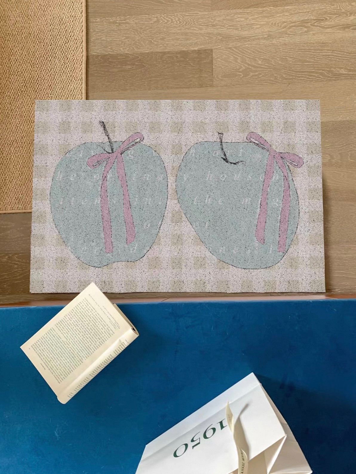 An apple doormat lies at a doorway with two green apples and pink bows over a pale yellow gingham pattern.