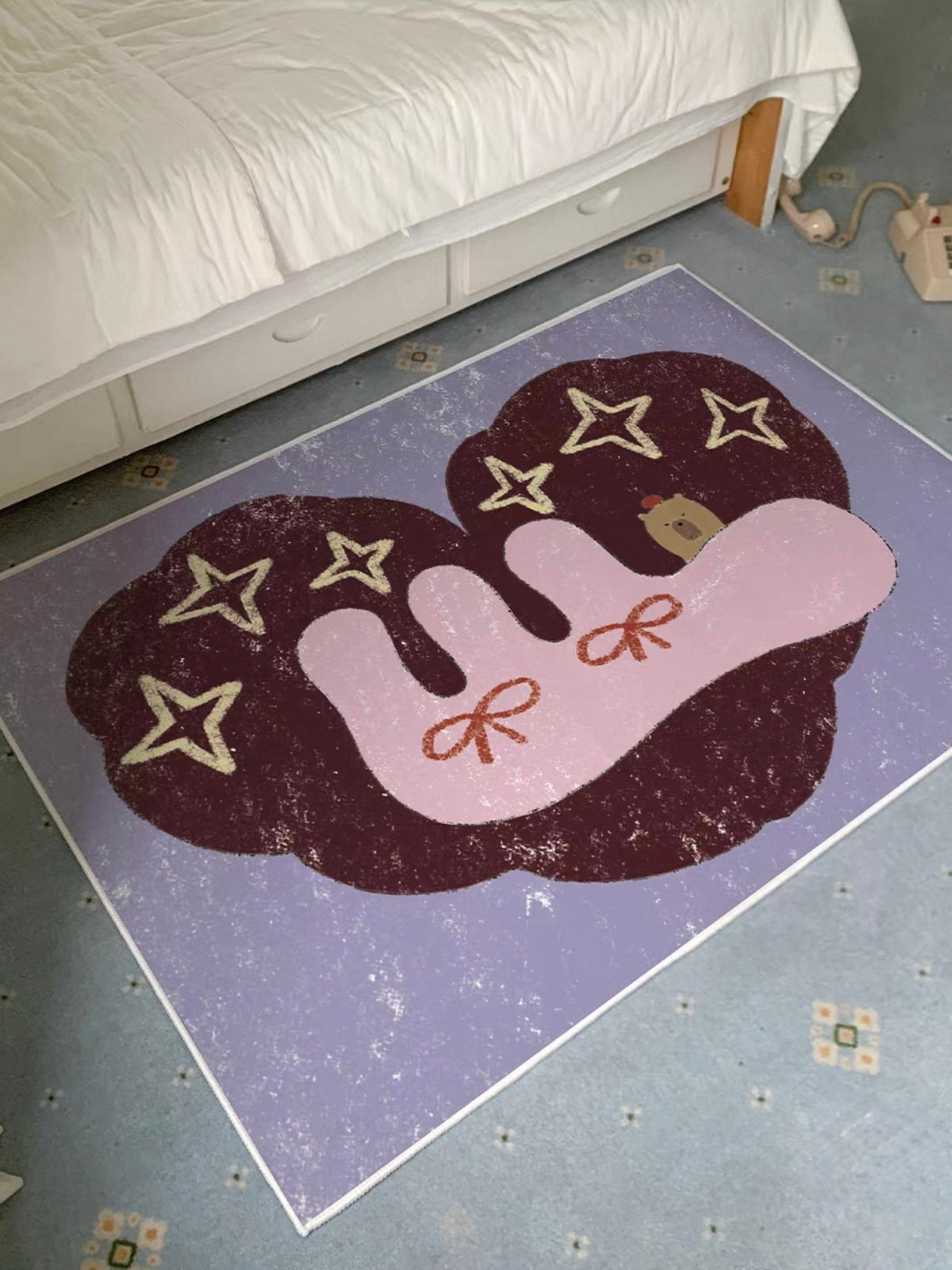 A bedside rug with pink bow details and pale stars rests beside a light bed on a purple rectangle.