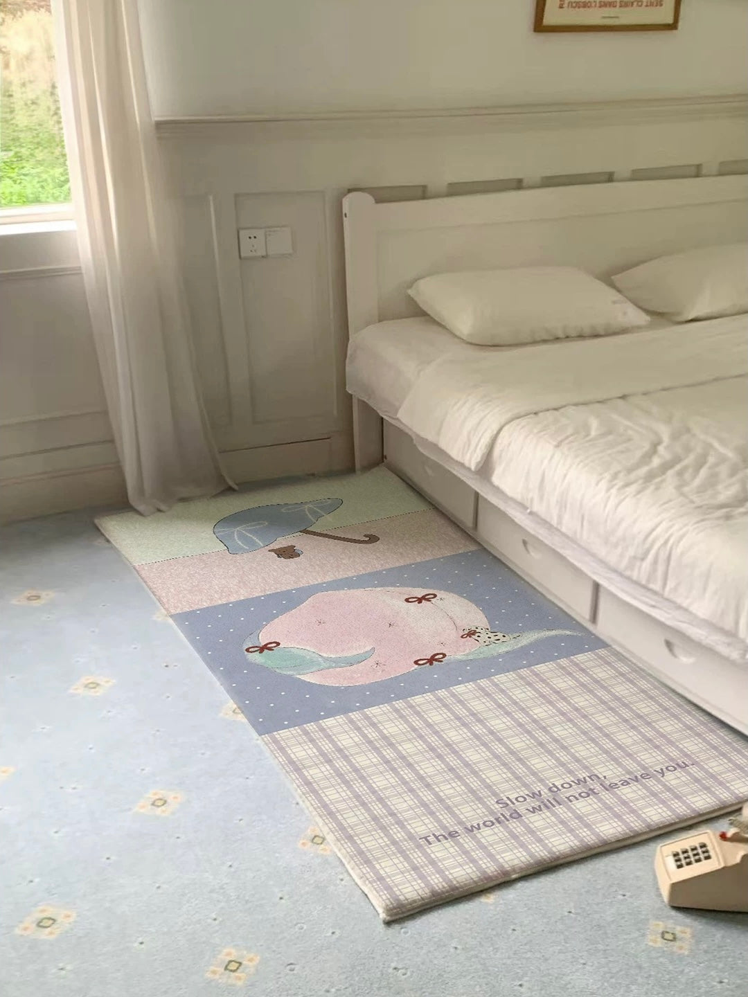 A bedside runner rug in lilac, pink and cream lies beside a bed, with a peach panel and checked section.