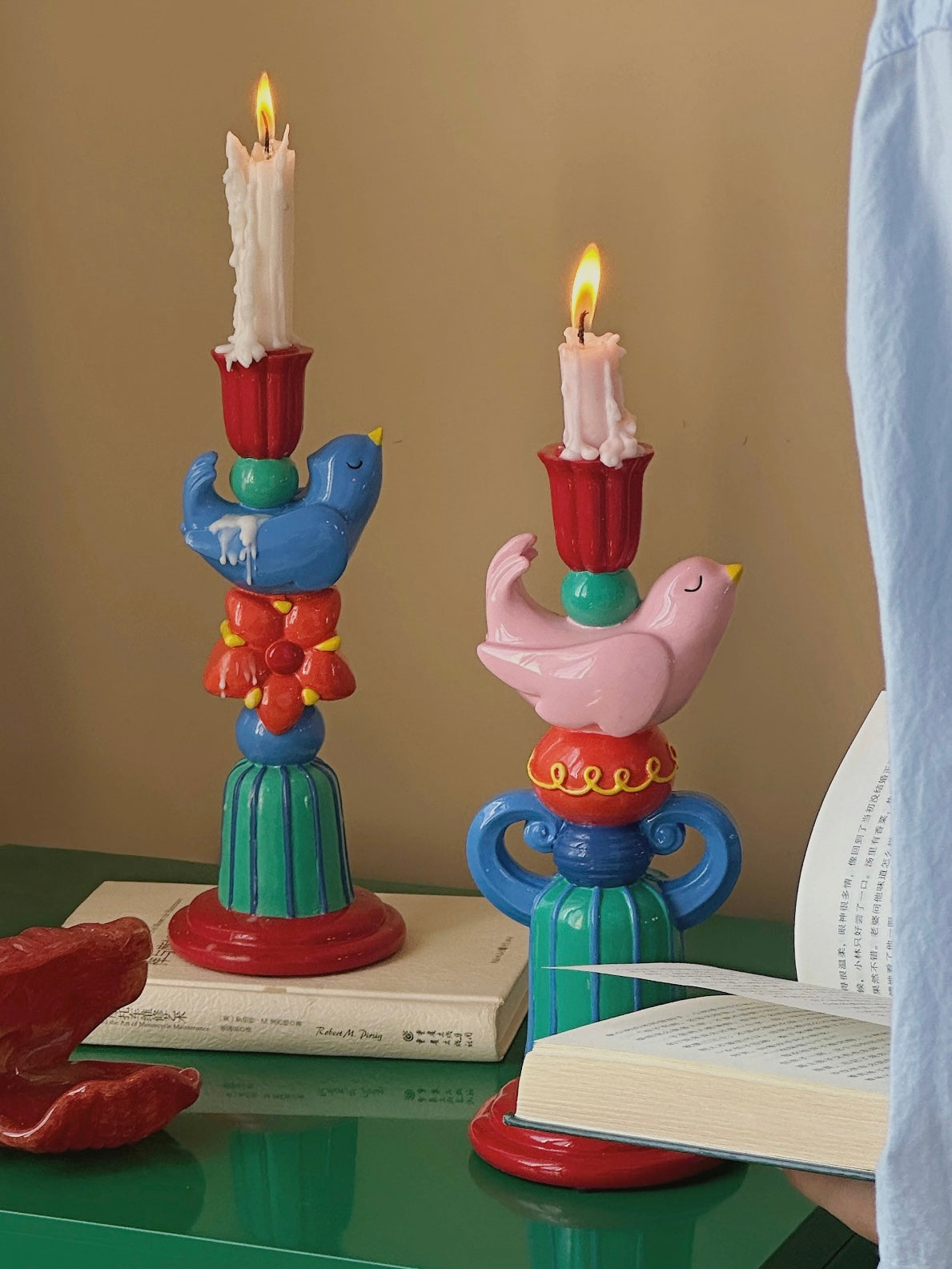 Pink and blue bird candle holders displayed with taper candles beside colorful wall art and leafy plants.
