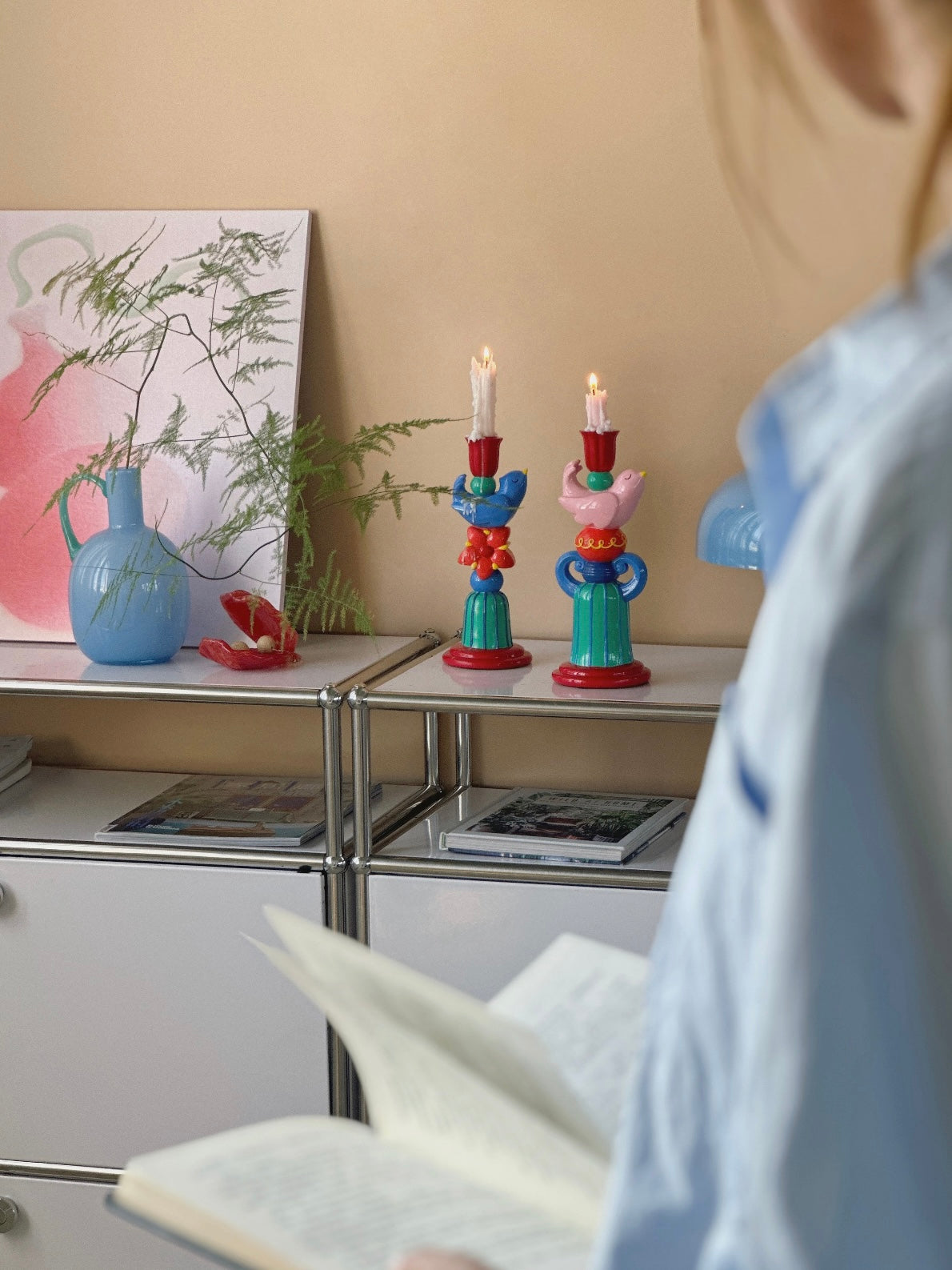 Pink and blue bird candle holders with lit candles displayed on a mirrored console beside a blue lamp.