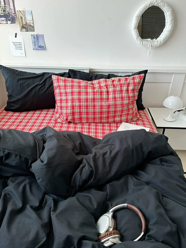 Unmade bed with red checkered blanket and black pillows, with a side table and mirror in the background.