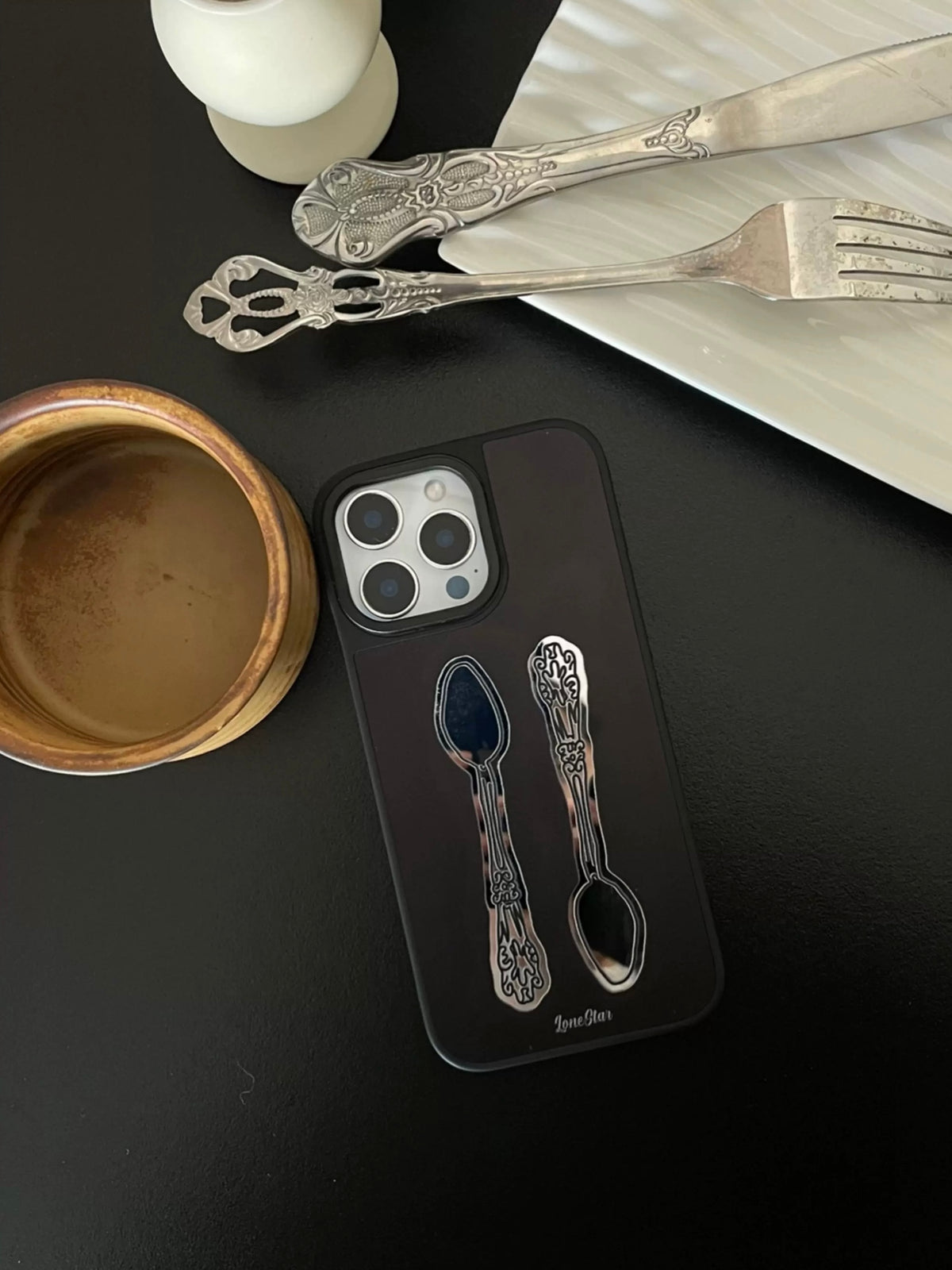 A black mirror phone case lies flat with two ornate spoon designs and a large square camera opening.