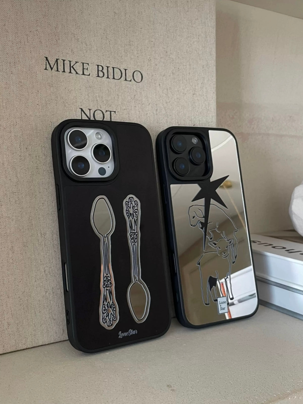 Black and silver phone cases stand side by side, displaying two vintage spoons and lamb outlines below a star.