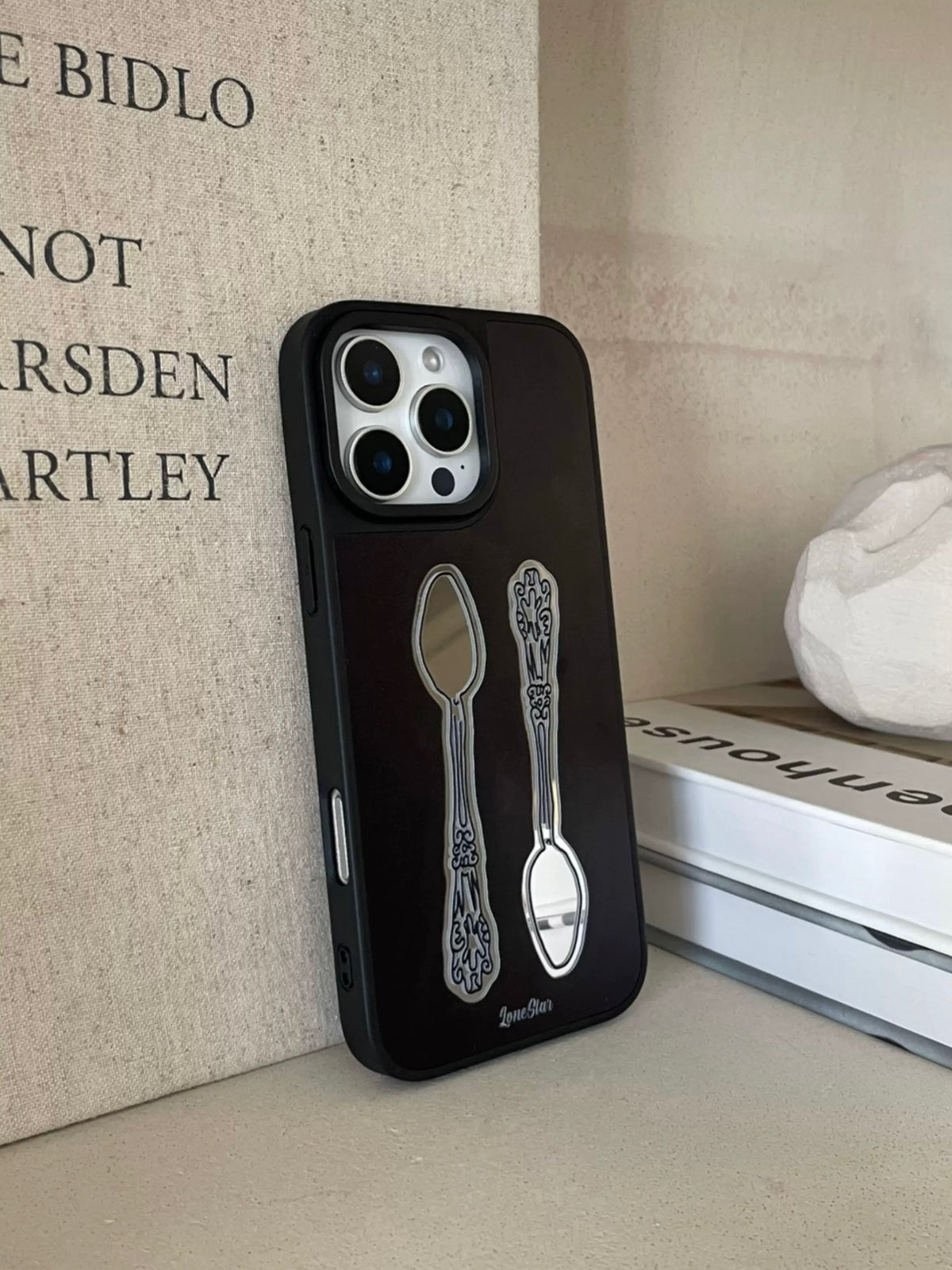 A black spoon phone case stands upright, showing two ornate spoon illustrations below the square camera opening.