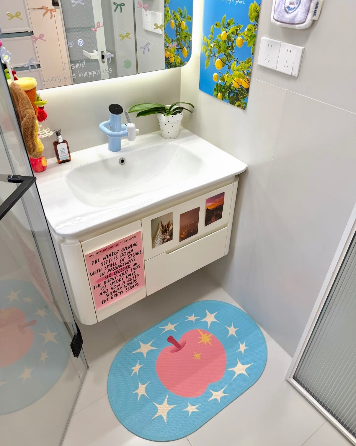 A bathroom vanity area with a white sink cabinet, mirror, wall art, and a blue oval floor mat featuring a pink apple and white stars. A glass shower door is visible on the left.