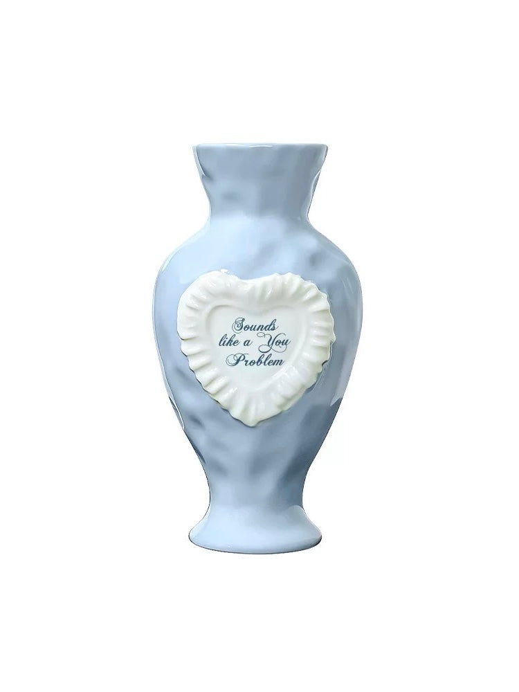 Blue ceramic vase with a raised white heart reading Sounds like a You Problem, a flared neck and rounded shoulders.