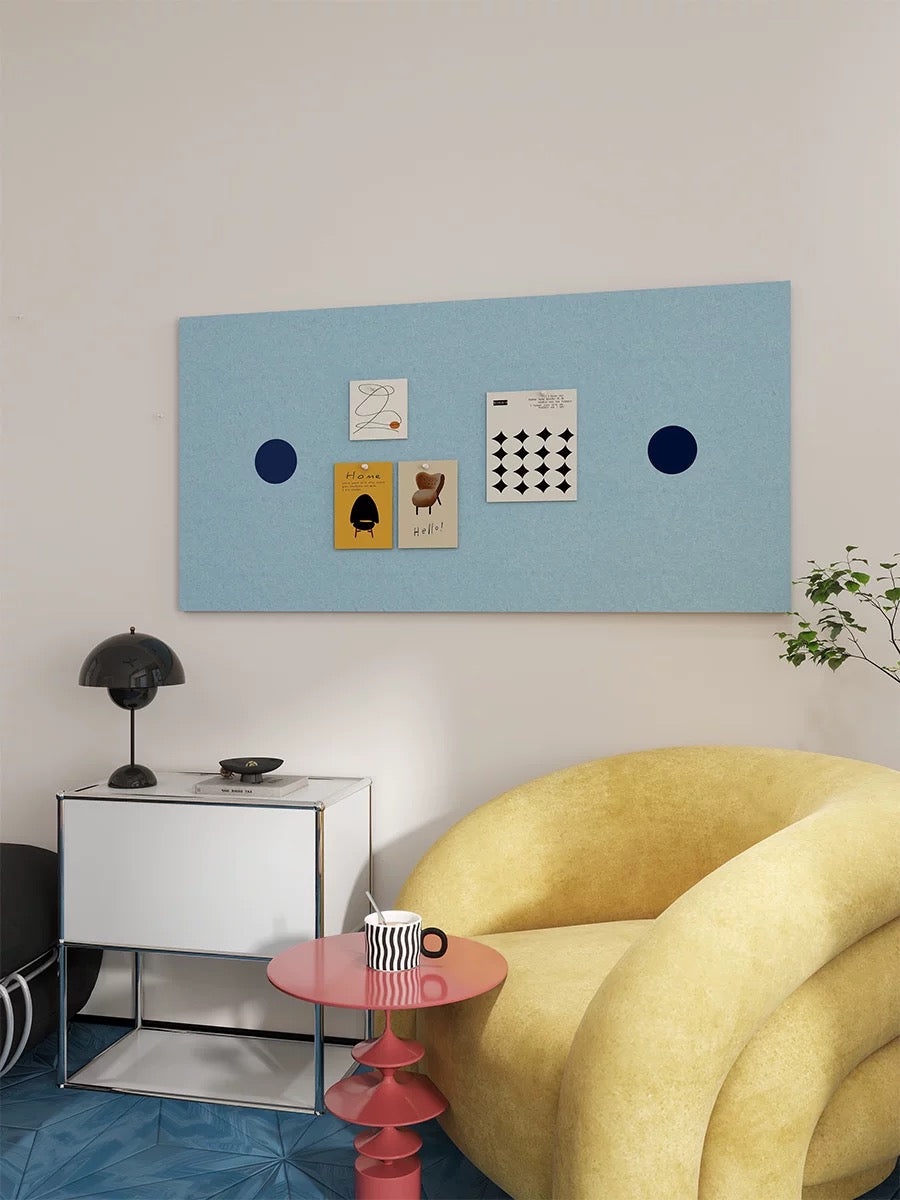 Blue felt pin board with navy circles and pinned art in a living room with a yellow chair.
