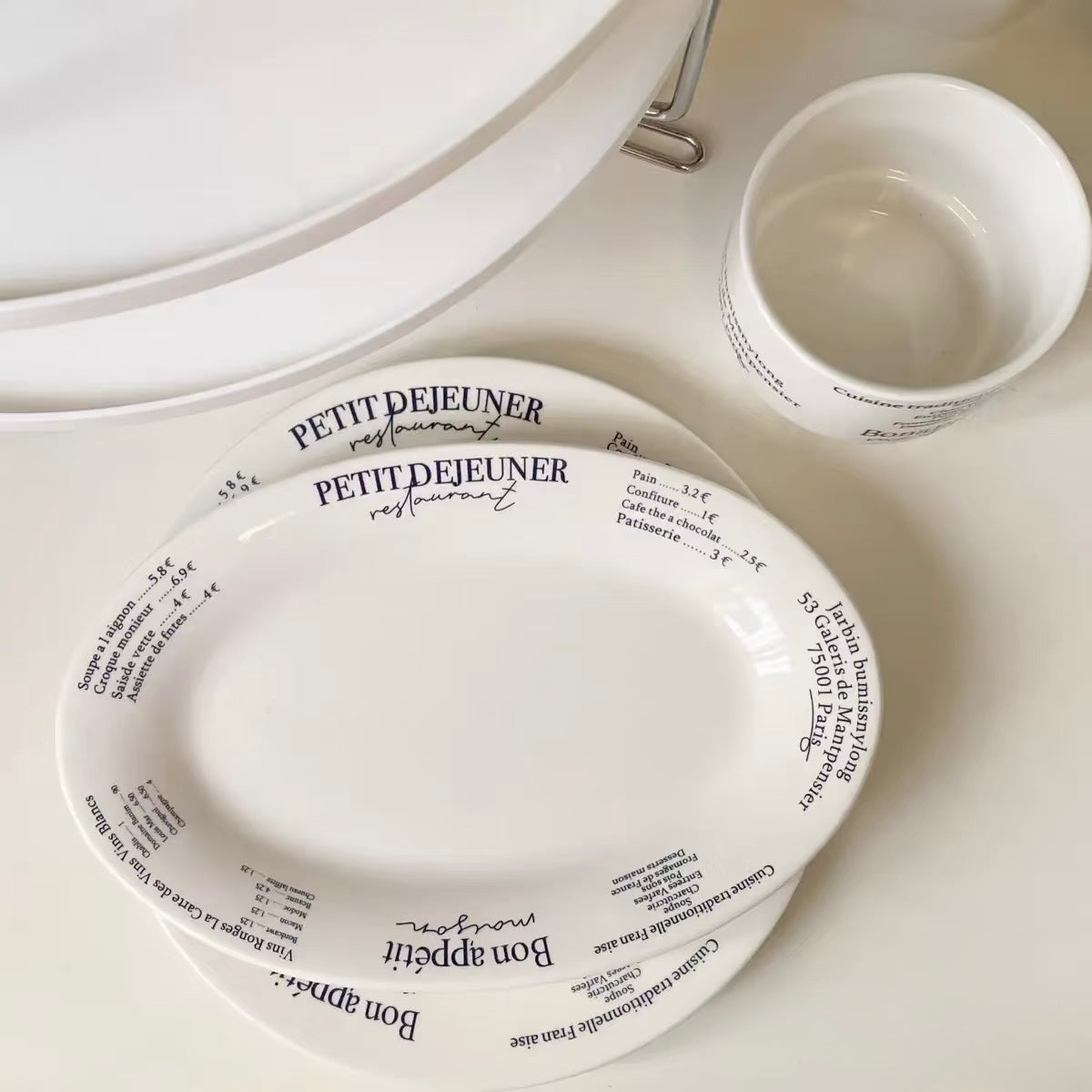 White ceramic tableware set featuring a platter with black printed French text, a matching cup, and stacked plates on a white surface.