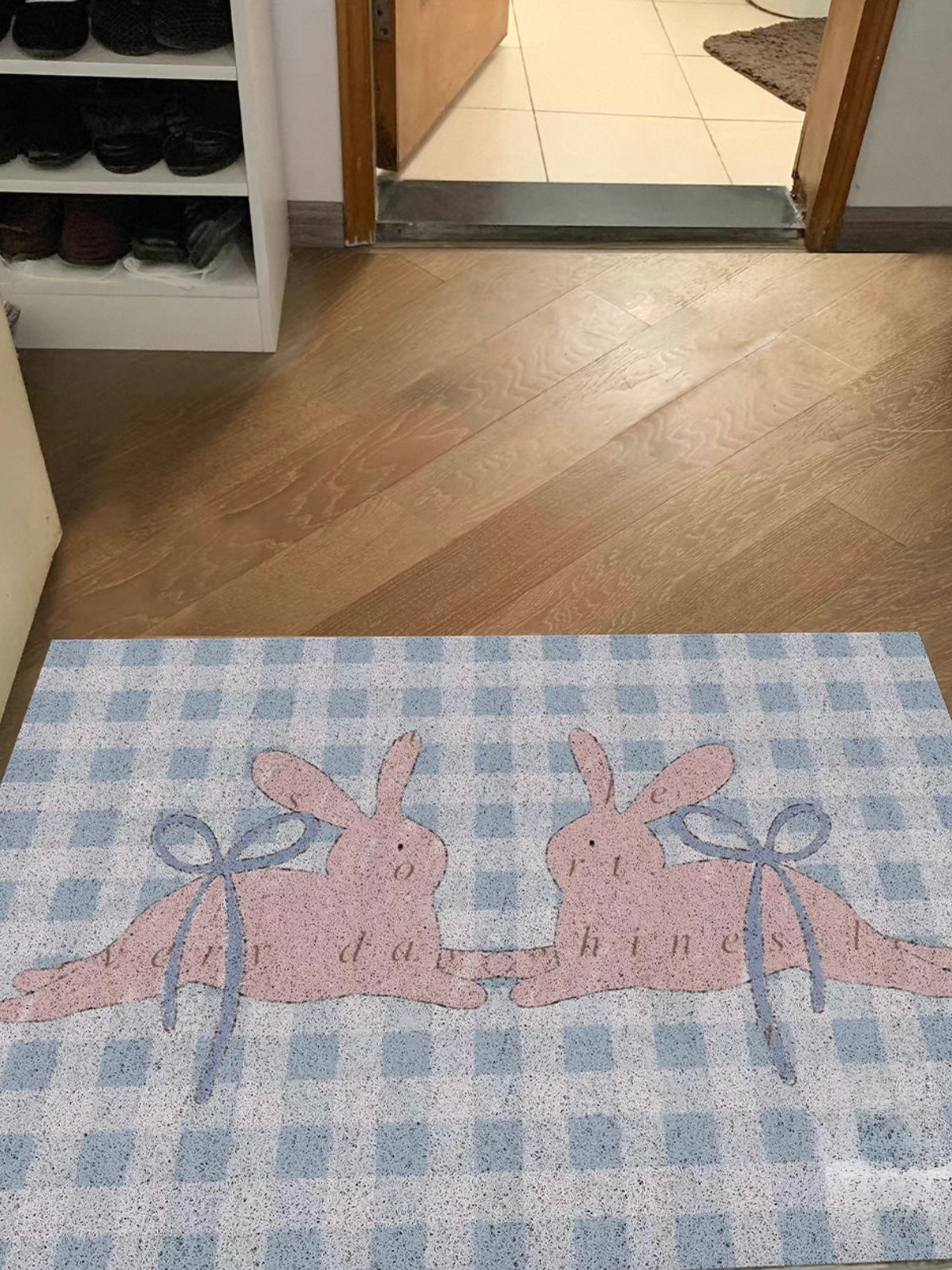 A blue gingham doormat sits at an open doorway with two pink bunnies and blue ribbon bow details.