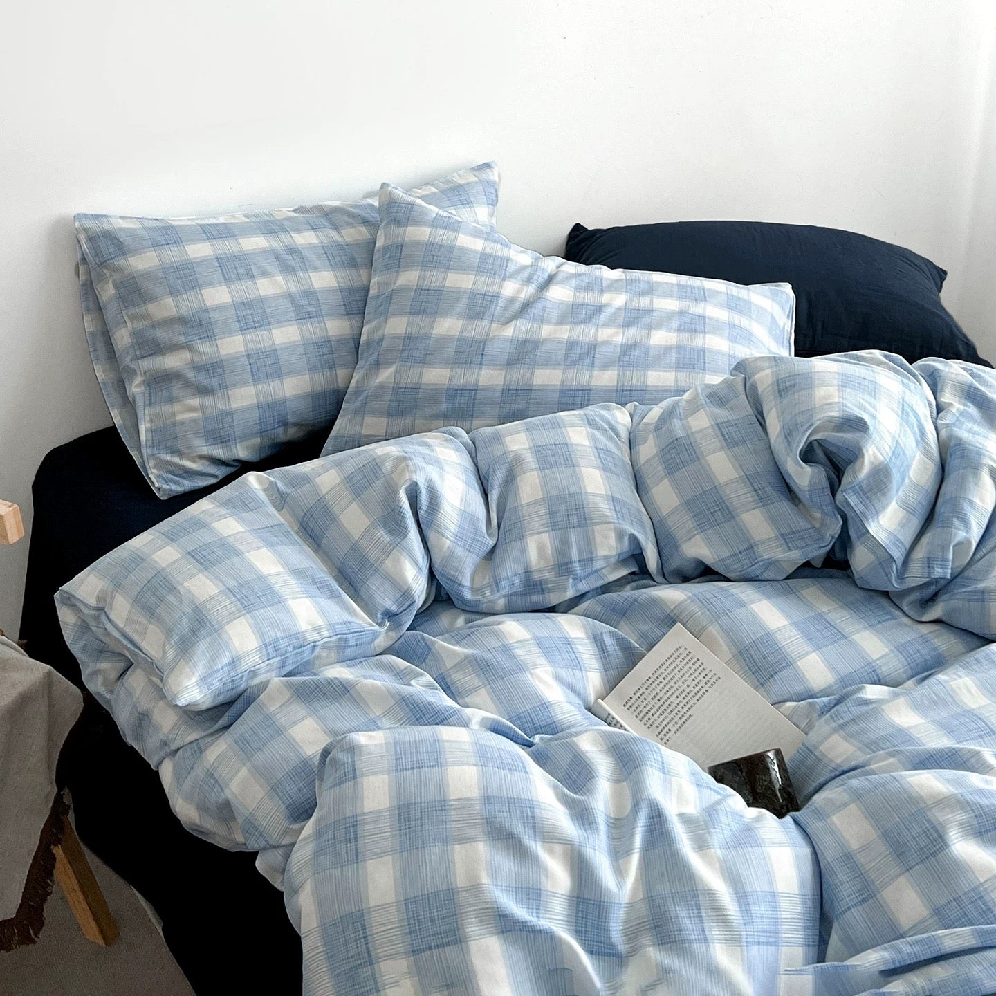 Light blue and white plaid bedding set with matching pillowcases and a duvet cover, styled on a bed with dark pillows and a book resting on the duvet.