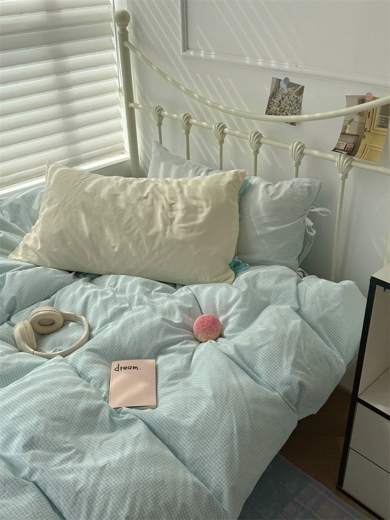 Bed with light blue bedding, white pillows, and a pink pom-pom on a white wall background.