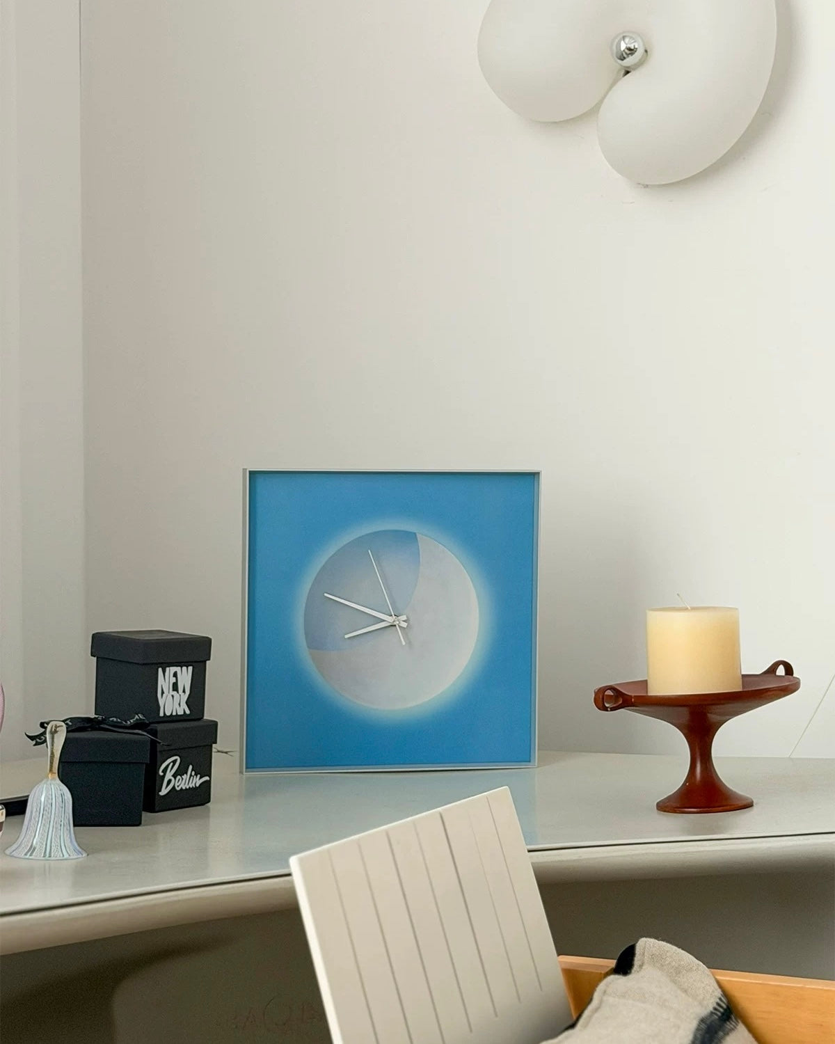 A square framed wall clock with a blue border and a minimalist white circular face sits on a light gray console table in a modern room setting, styled with a candle, decorative boxes, and a chair.