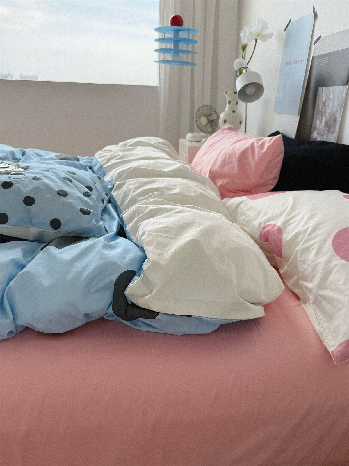 A view from the window side shows a pink sheet, layered pink and black pillows, and blue dotted bedding with a pale folded section.