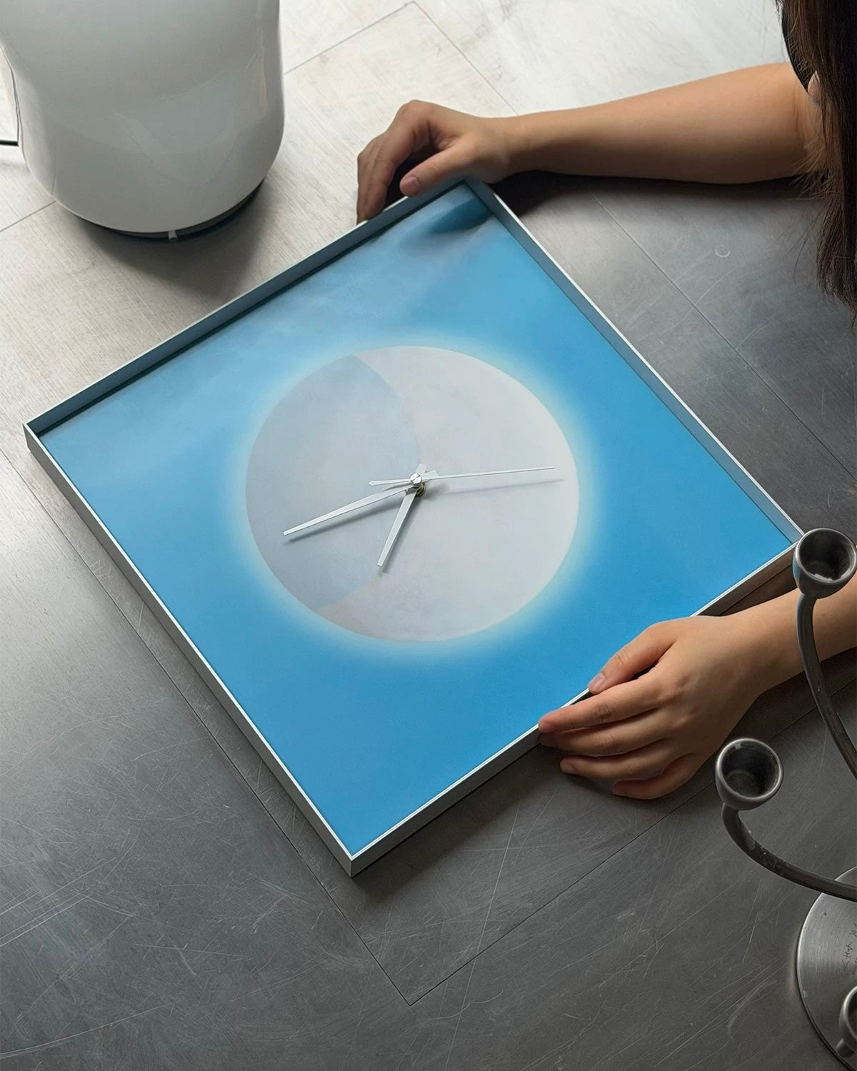 A square wall clock with a blue and white circular design, held by a person's hands on a gray tiled floor.