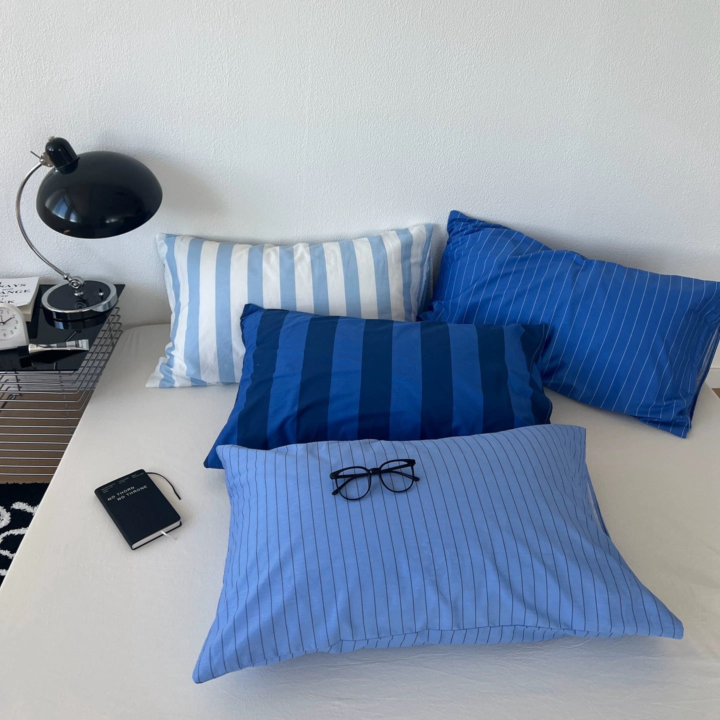 Three blue and white striped and pinstriped pillows arranged on a bed next to a black desk lamp and notebook.