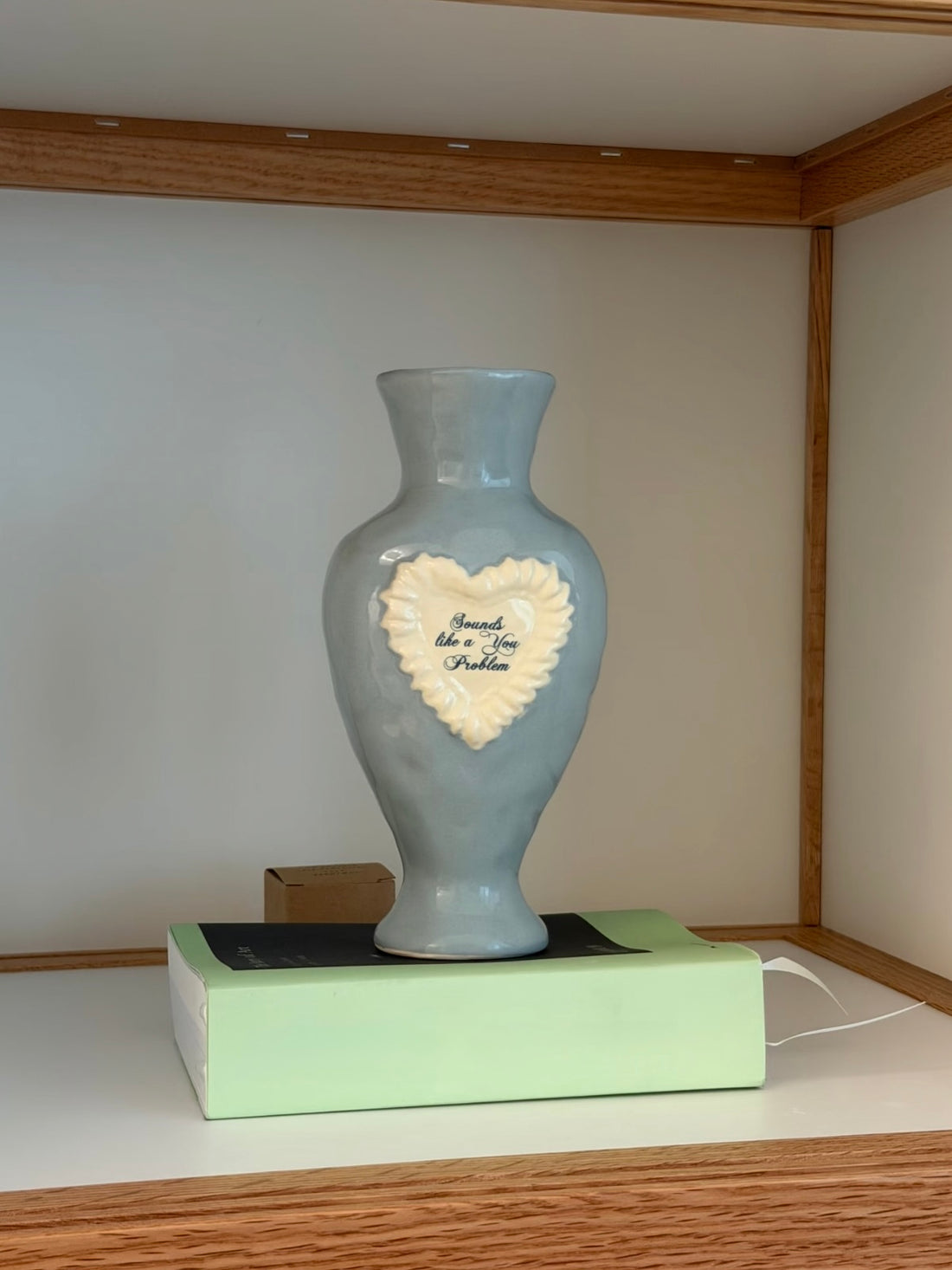 Blue vase standing on a pale green box, with a raised white heart, blue script and a flared foot below the body.
