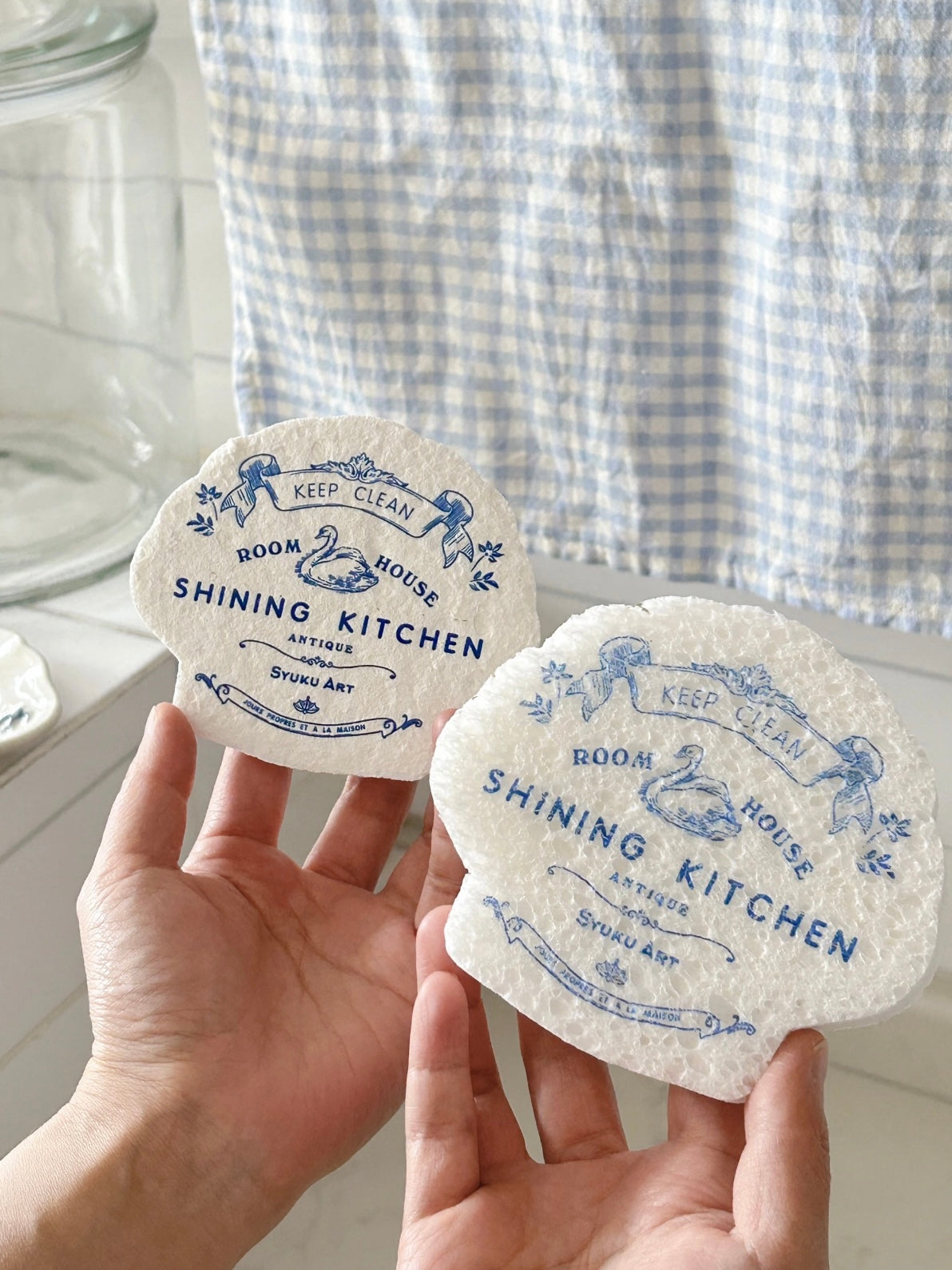 Two blue and white dish sponges are held side by side, showing scalloped edges and matching swan artwork.