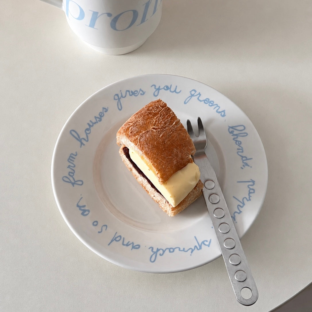 A white ceramic plate with blue cursive text, holding a poached egg with a drizzle of honey and black pepper, styled on top of a gray book with gold lettering on a white bed alongside a laptop and a cream-colored plate with a knife.