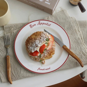 A white ceramic plate with red trim, printed with 'Bon Appetit' and 'French mood', holding a strawberry and cream cake. The plate is styled on a beige textured placemat with a wooden-handled knife, a fork, a cream mug, and a book in the background.