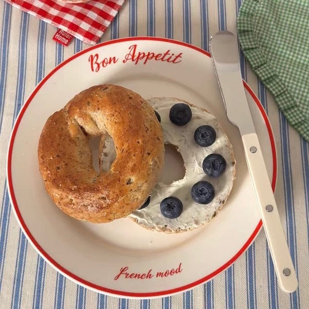 A white ceramic plate with red trim, printed with 'Bon Appetit' and 'French mood', holding a strawberry and cream cake. The plate is styled on a beige textured placemat with a wooden-handled knife, a fork, a cream mug, and a book in the background.