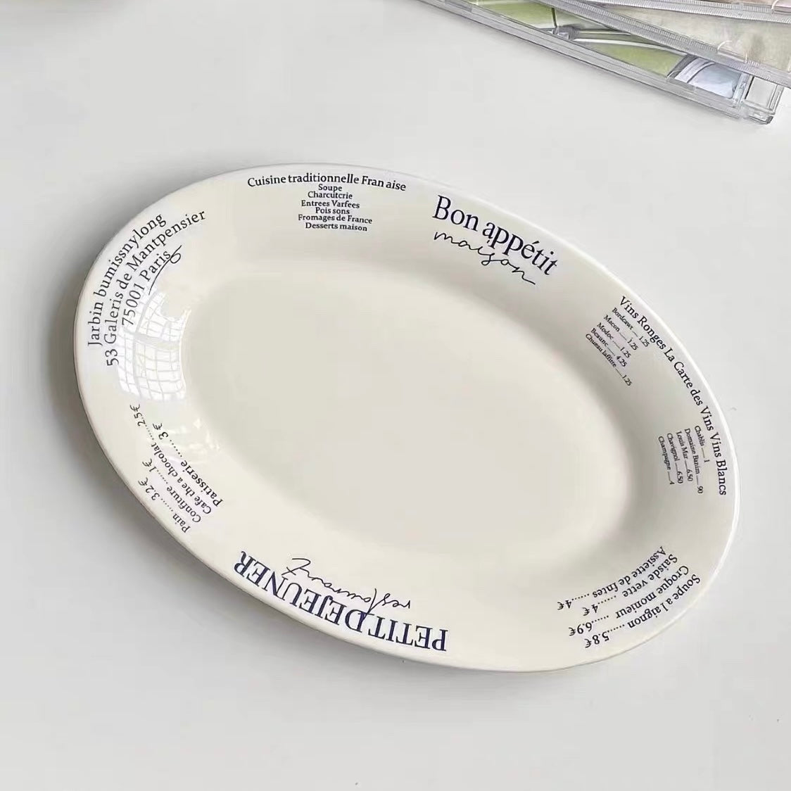 An oval white ceramic serving platter with blue French text and decorative script, photographed on a light gray surface with part of a clear plastic bag visible in the top right corner.