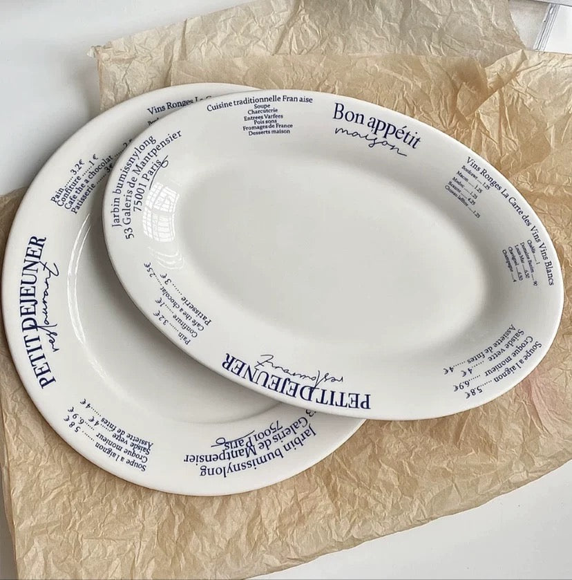 Two white oval serving platters with black printed French text and the name 'Petit Deleuner' around the rim, arranged on crumpled brown paper.