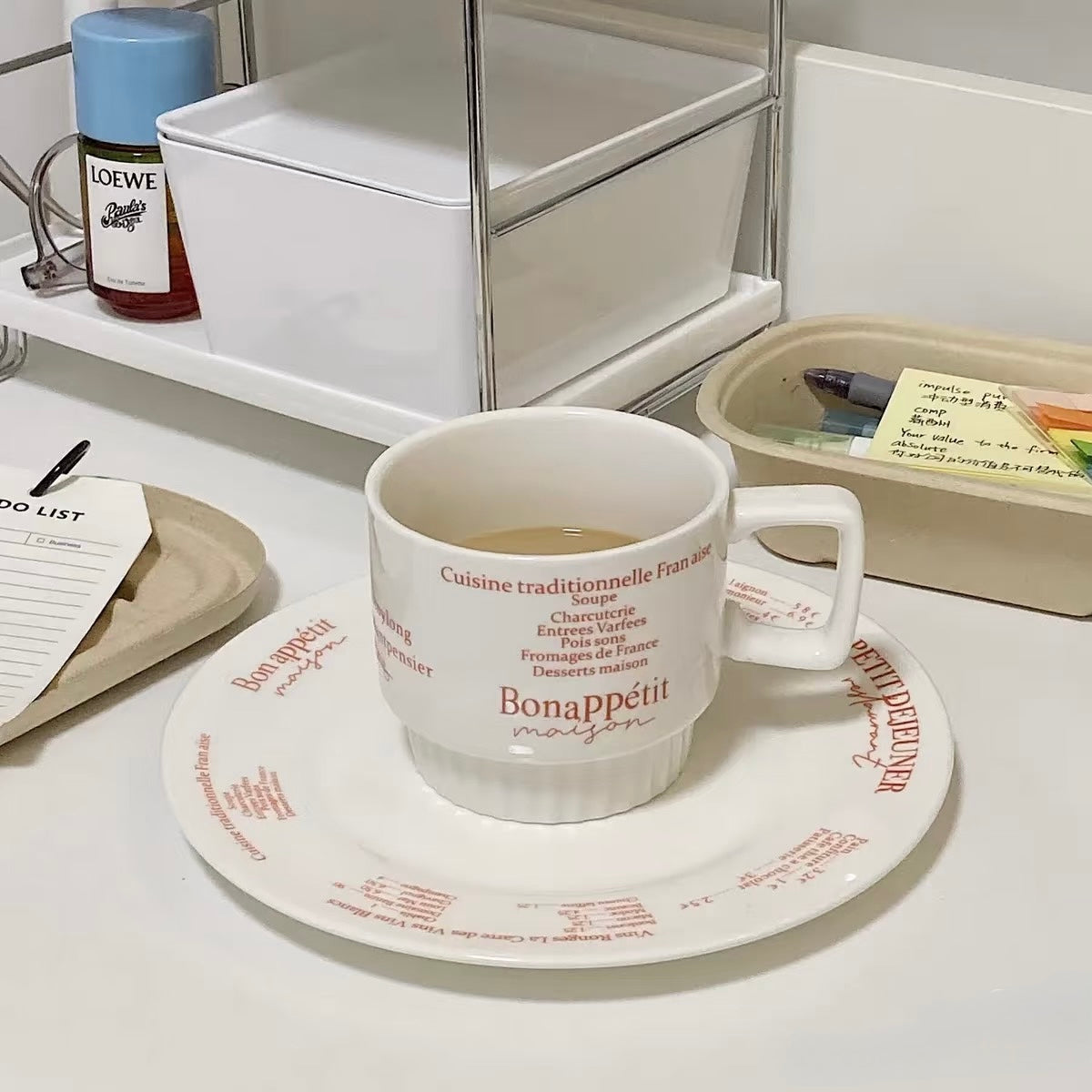 A white ceramic coffee cup with red French text reading 'Bonappetit maison' sits on a matching saucer in a styled tabletop setting. The scene includes a Loewe bottle, white storage bins, and a beige tray in the background.