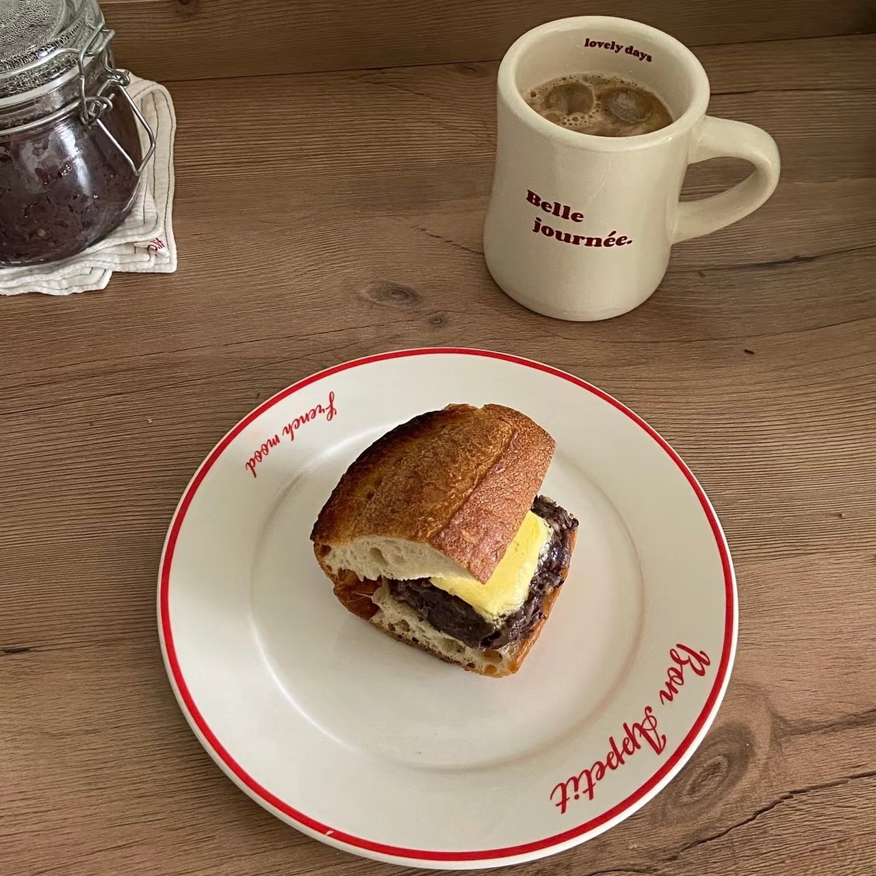 A breakfast scene on a wooden table with a plate holding a sandwich on a white plate with red trim, a cream-colored coffee mug with red text reading 'elle journée.', and a glass jar partially visible on the left.