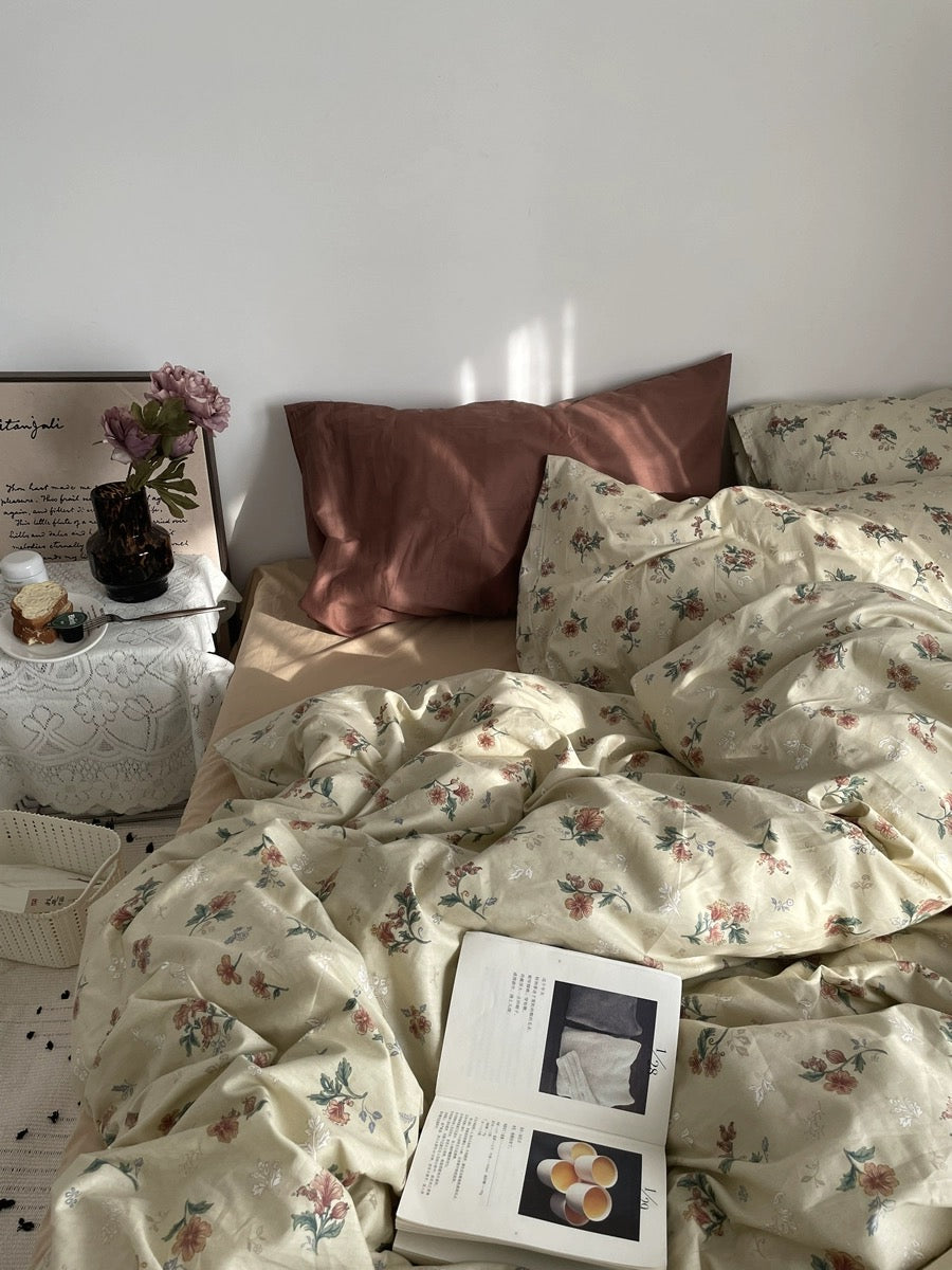 Vintage floral duvet on a sunlit bed with rust pillows, open book, flowers, and bedside table.