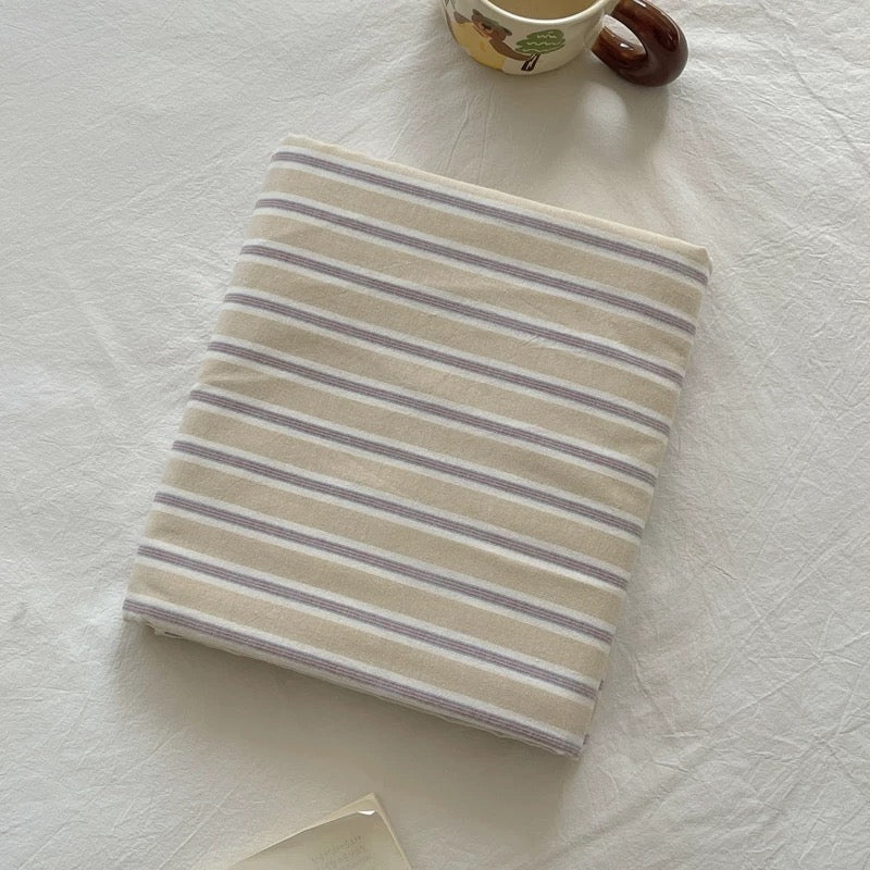 A folded cream-colored fabric item with thin horizontal gray stripes, placed on a white textured fabric surface with a small decorative mug partially visible at the top.