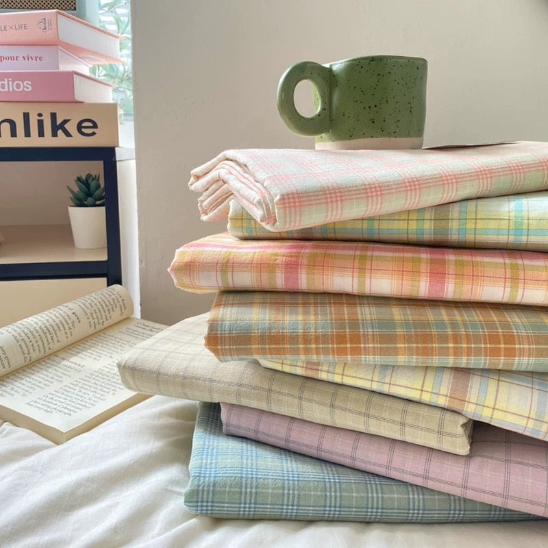 A stack of folded plaid fabric sheets or blankets in pastel colors, arranged on a bed with a green speckled mug on top. An open book and a small side table with books are visible in the background.