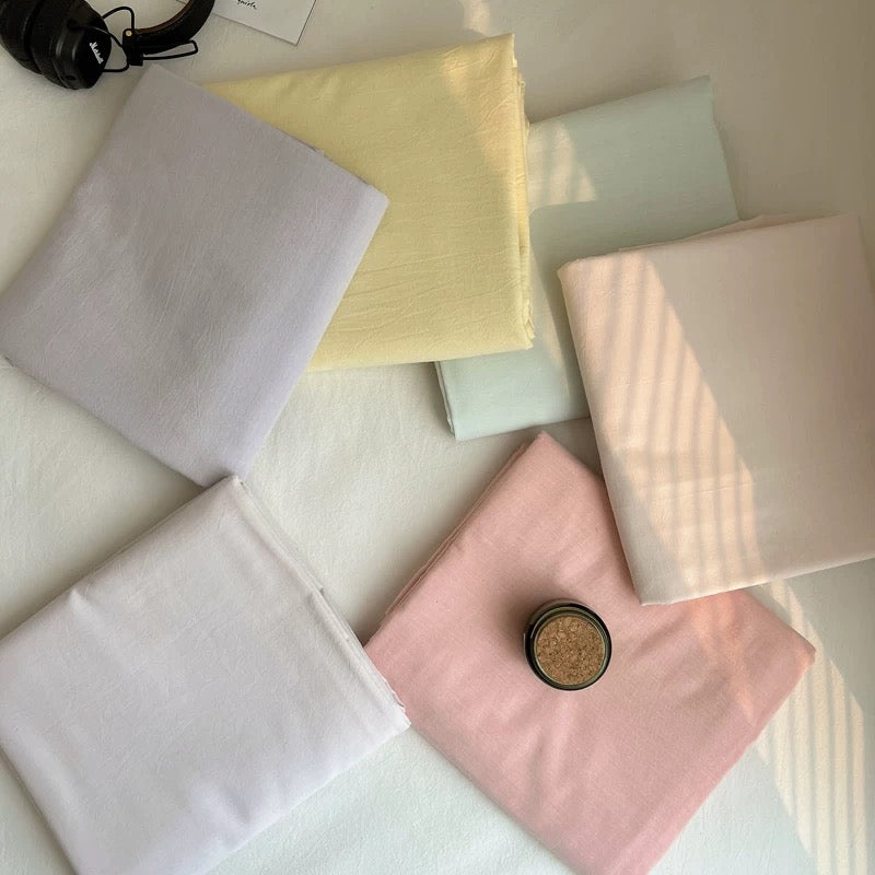 Six folded fabric swatches in pastel shades of lavender, pale yellow, mint, blush pink, cream, and white, arranged on a light surface with a small round jar on the pink fabric.