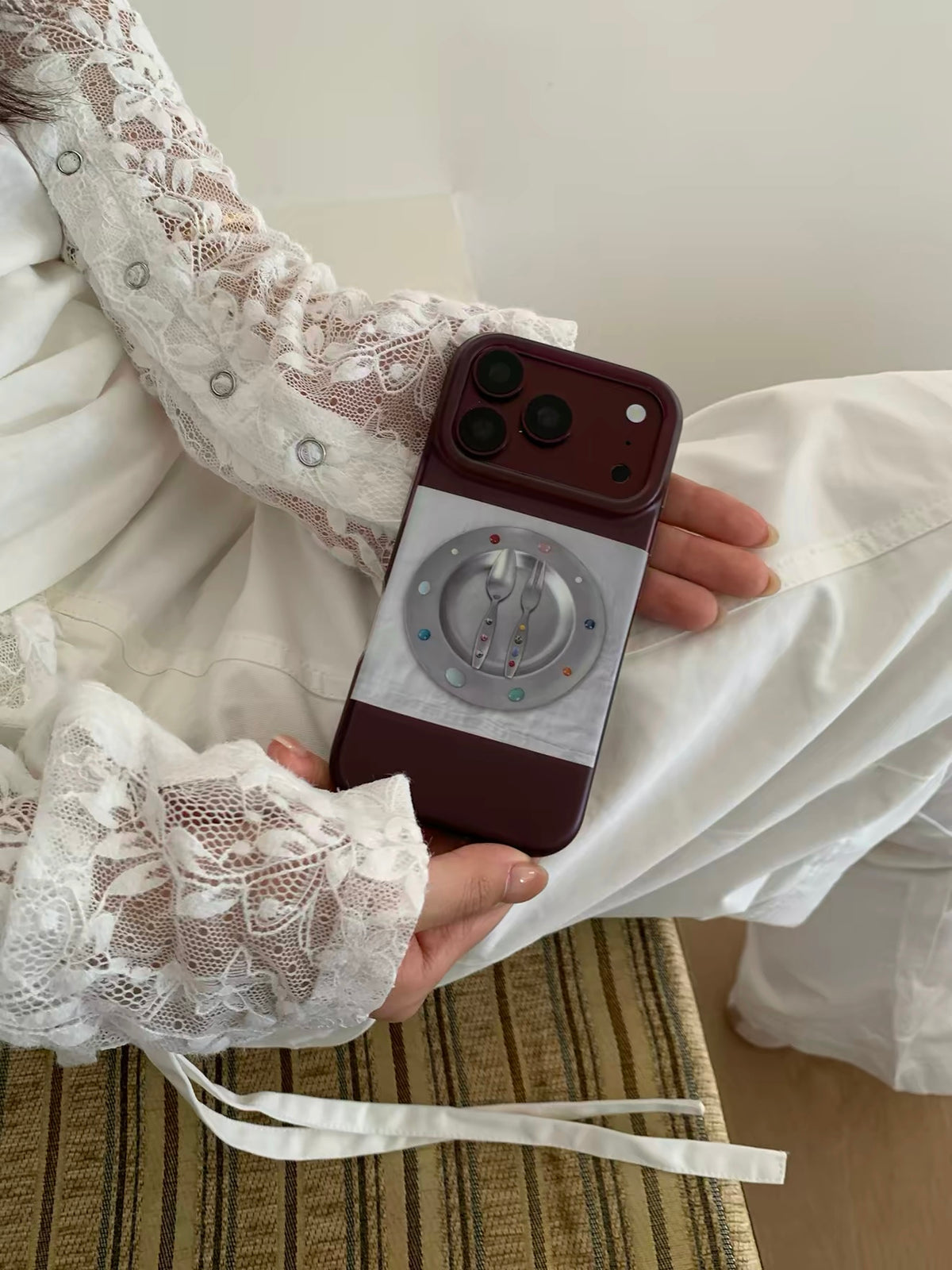 A burgundy iPhone case held upright in one hand, showing a pale plate and cutlery design across the back.