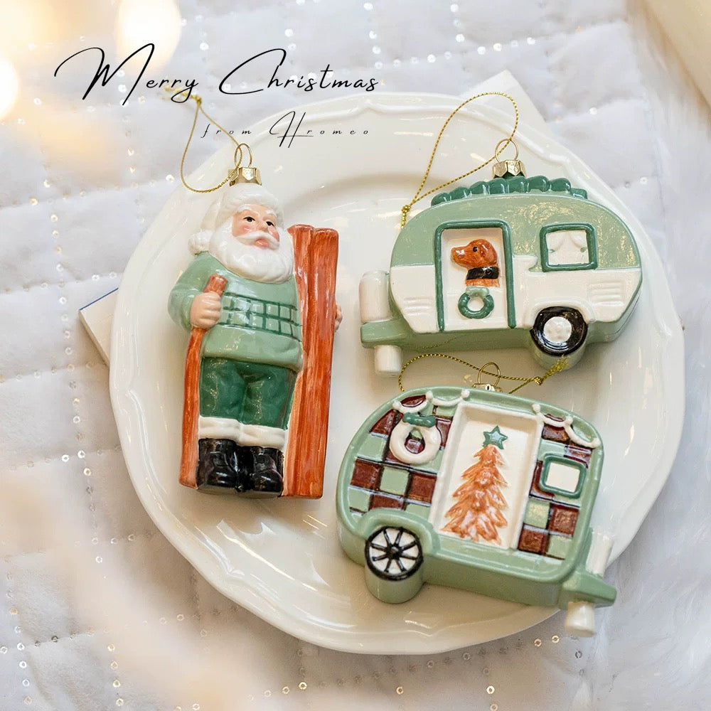 Camper ornaments with a tree panel and a dog at the door sit beside a Santa figure in a green sweater.
