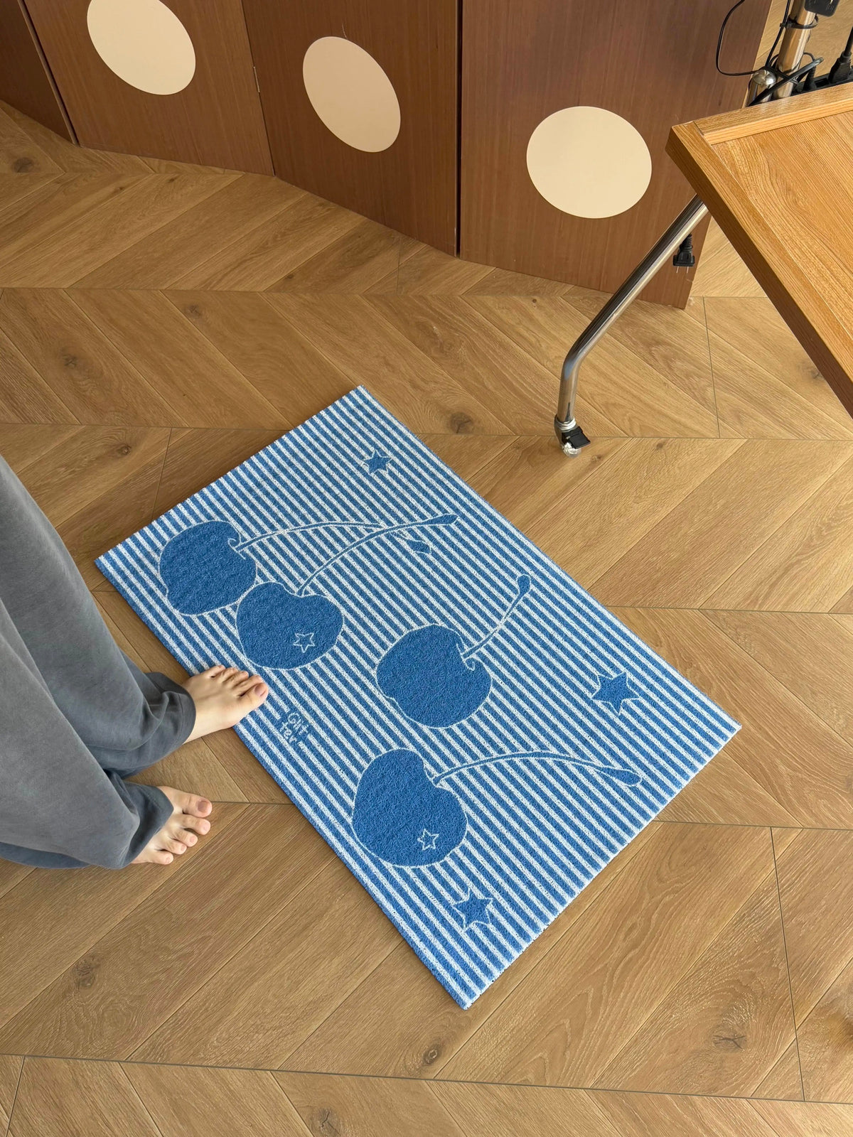 A blue and white striped rectangular mat with a cherry pattern lies on a light wood herringbone floor. A person's bare foot and gray pants are visible at the left edge, and part of a wooden table and cabinet is visible in the room setting.