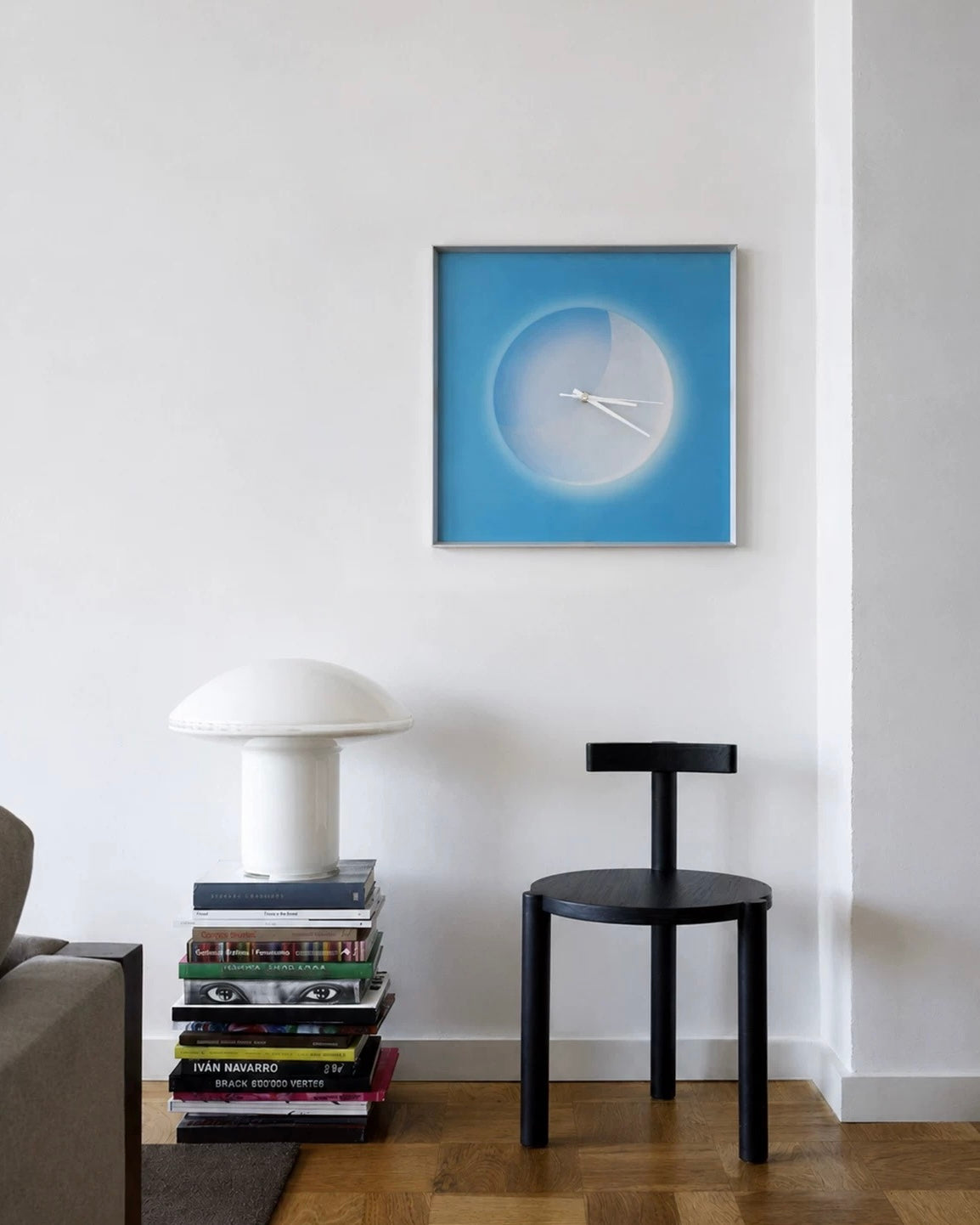 A square framed wall clock with a blue and white abstract design hangs on a white wall in a modern room setting, next to a black stool and a stack of books with a white mushroom lamp on top.