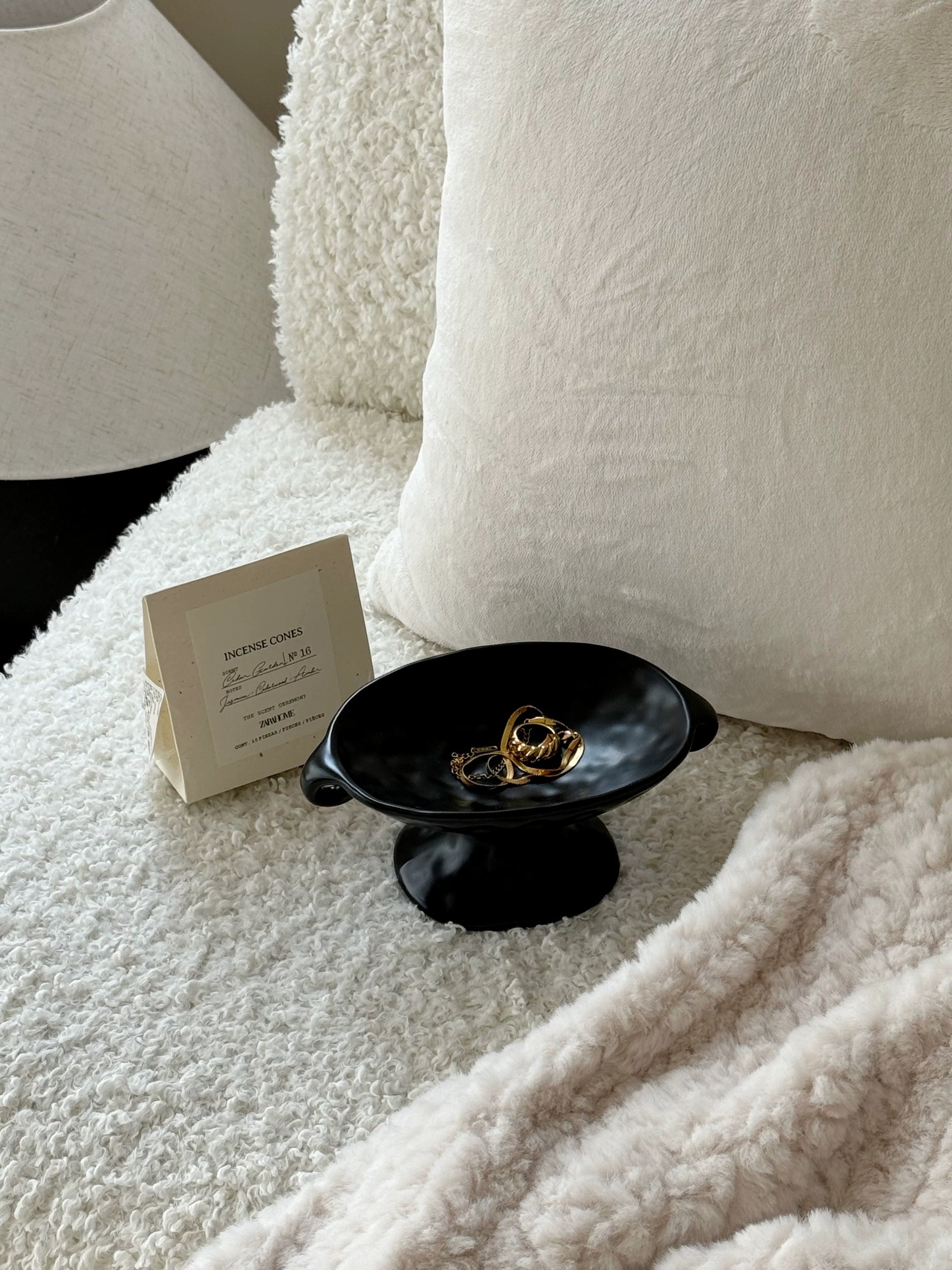 A small black pedestal bowl holding gold rings, styled on a white textured blanket beside a cream card and soft white pillows.