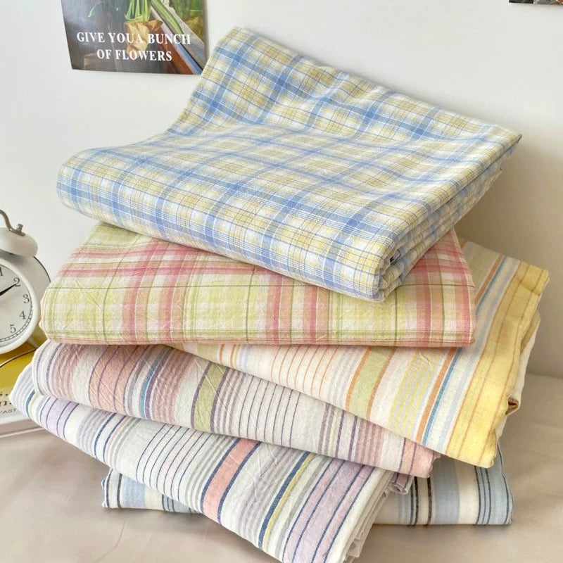 Four folded plaid fabric squares stacked on a light surface, each showing a different pastel plaid pattern: blue and yellow, mint and peach, pink and purple, and coral and cream. A dark green candle jar sits on the top folded fabric.