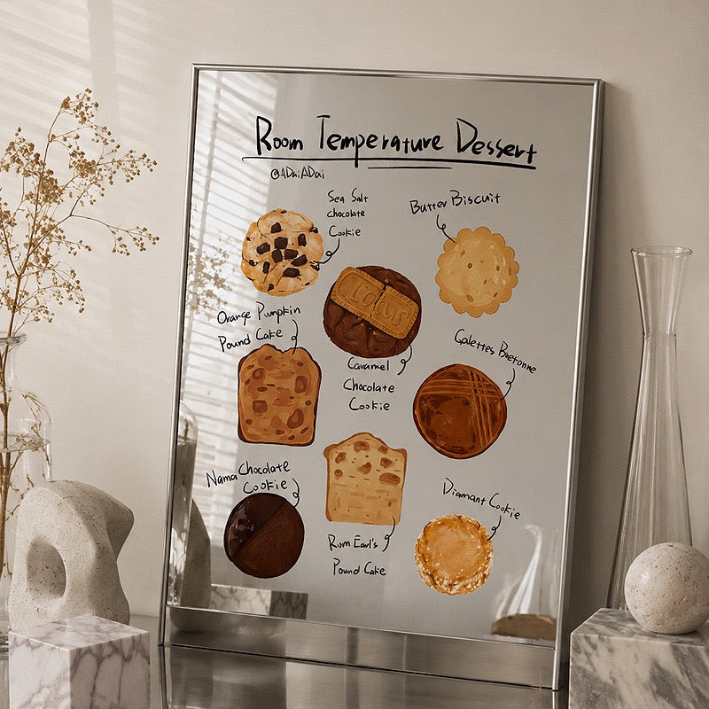 A framed art print titled 'Room Temperature Dessert' featuring illustrated cookies and cakes with handwritten labels, displayed on a marble surface in a styled interior setting.