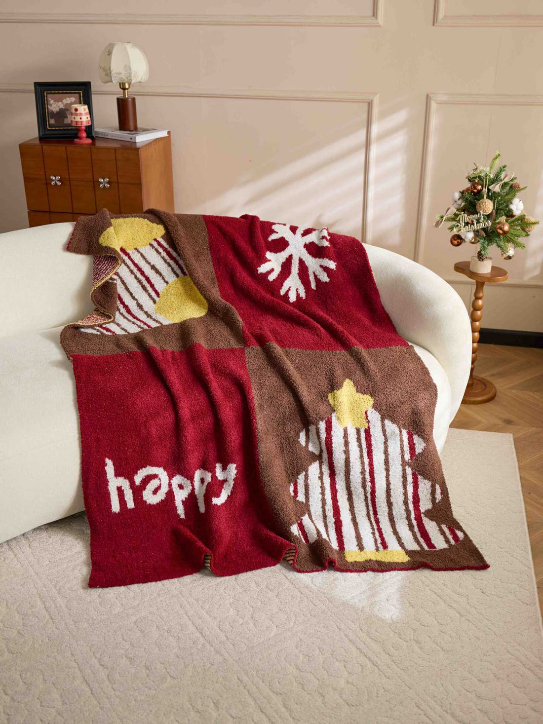 A person holds up a large red and brown holiday-themed blanket in a bright living room. The blanket has a four-quadrant design with Christmas tree, snowflake, striped ornament, and 'happy' motifs in red, brown, yellow, and white.