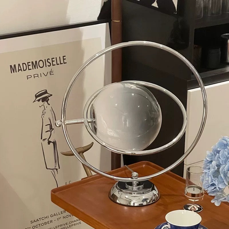 A modern table lamp with a spherical frosted white glass shade and a chrome metal frame, resting on an orange side table against a light wall.