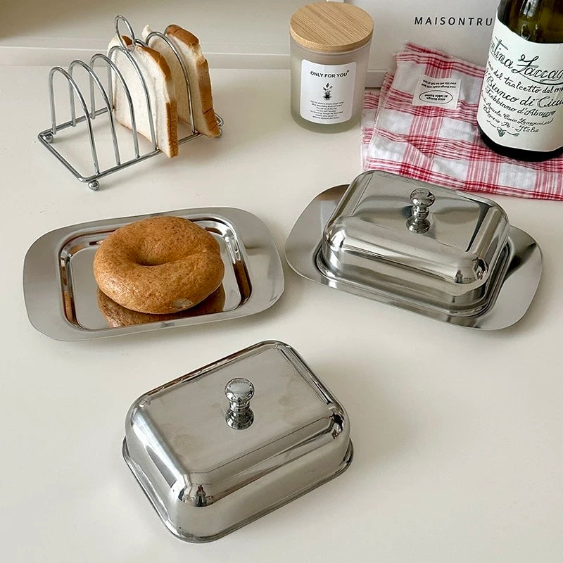 Silver butter dish with a lid, a donut, and a toaster rack with bread on a white surface.