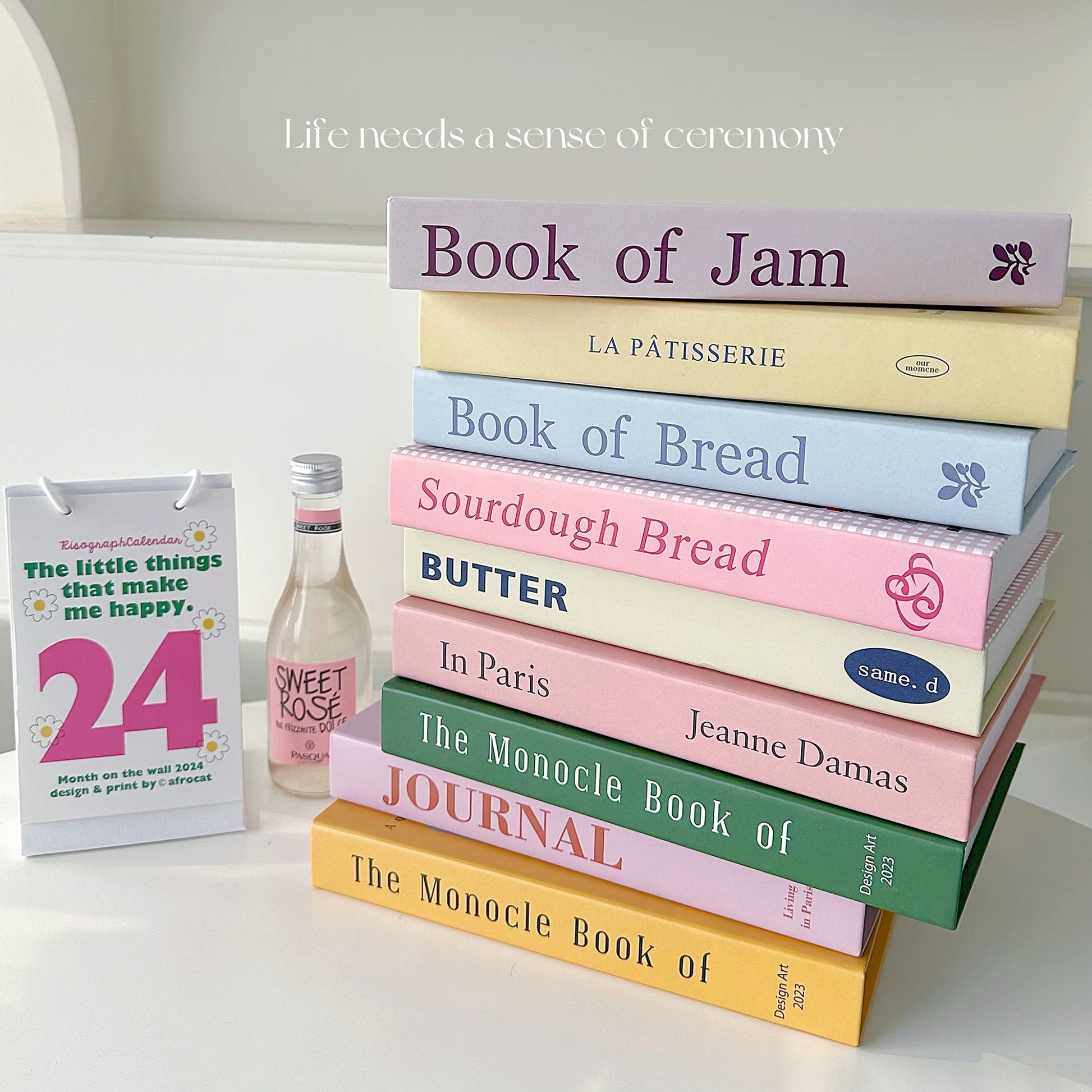 Stack of books with colorful covers on a white surface, with a bottle and calendar in the background.