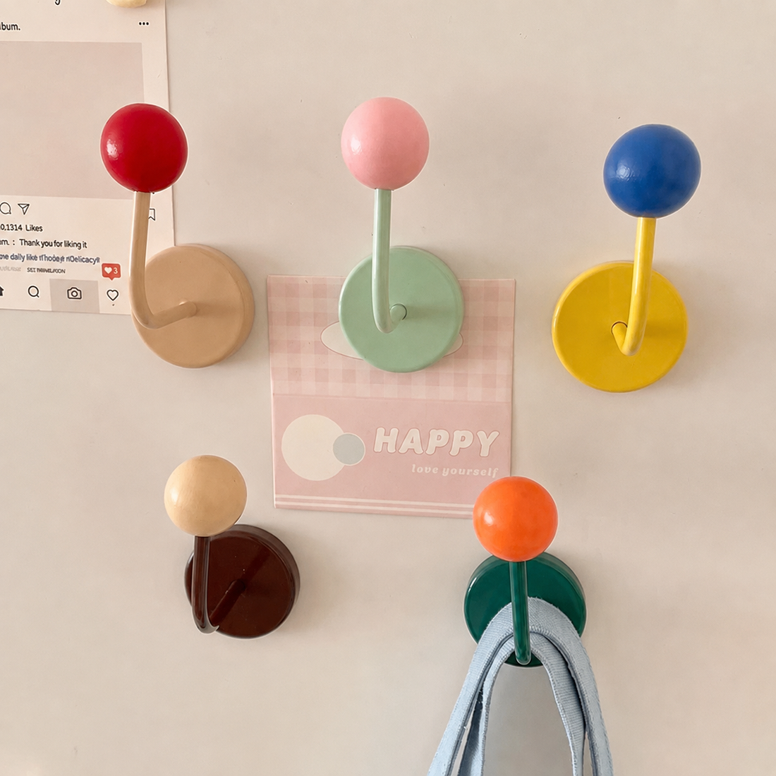 A set of six wall-mounted coat racks with round, colorful knobs and curved hooks, shown hanging on a white wall next to a printed Instagram post.