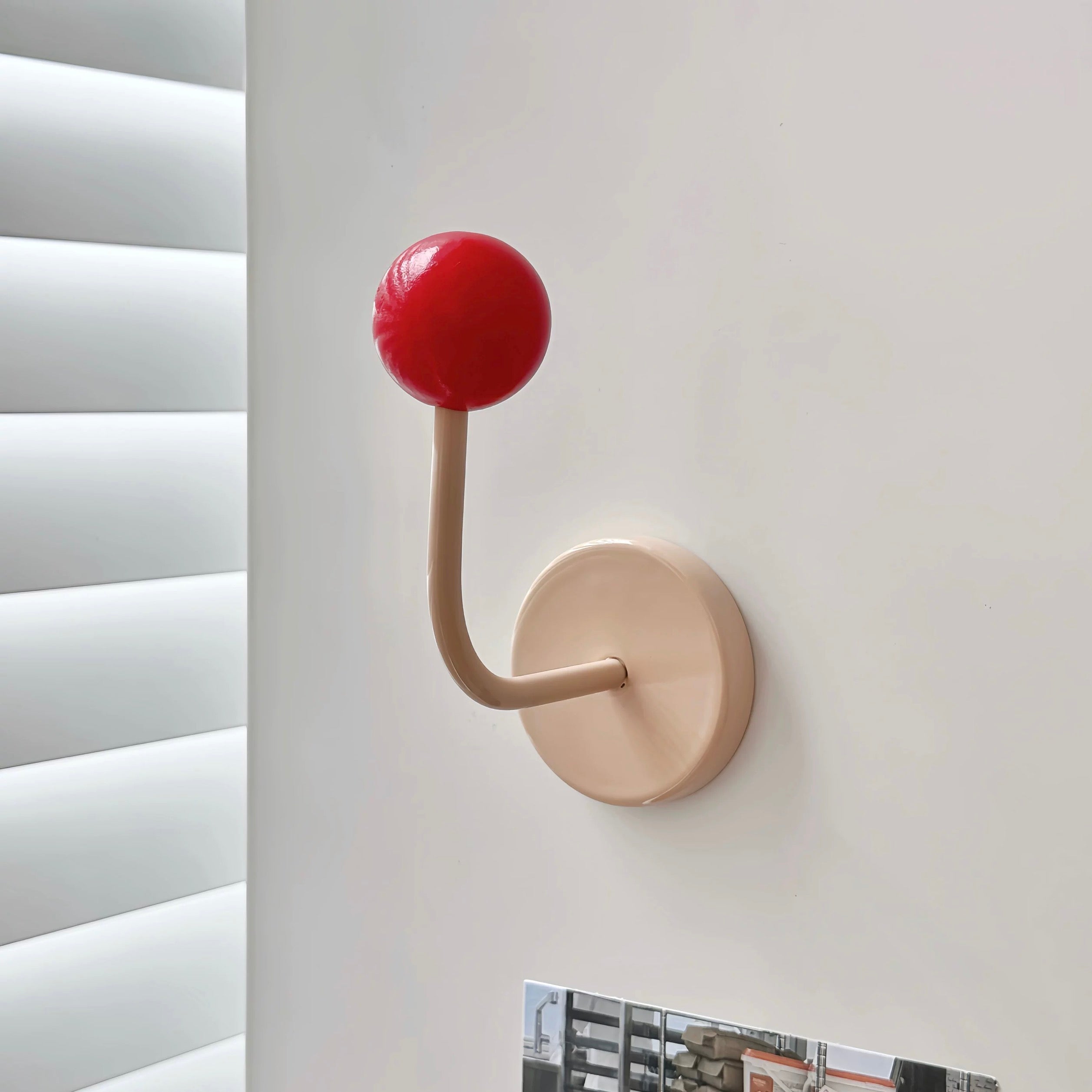 A minimalist wall hook with a round red top and a beige curved stem mounted on a beige circular base, shown on a white wall next to window blinds.